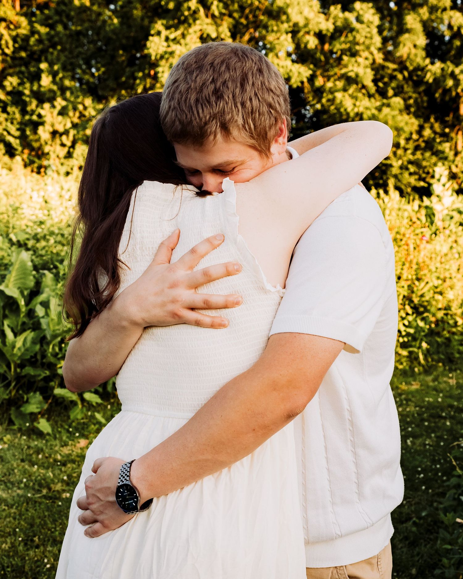 A couple shares a tender embrace outdoors, surrounded by lush green foliage in warm golden sunlight.