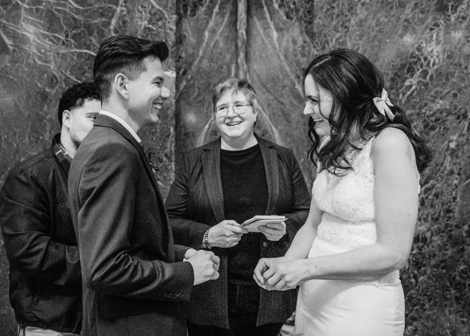 Black and white photo of bride and groom exchanging rings with smiling officiant.
