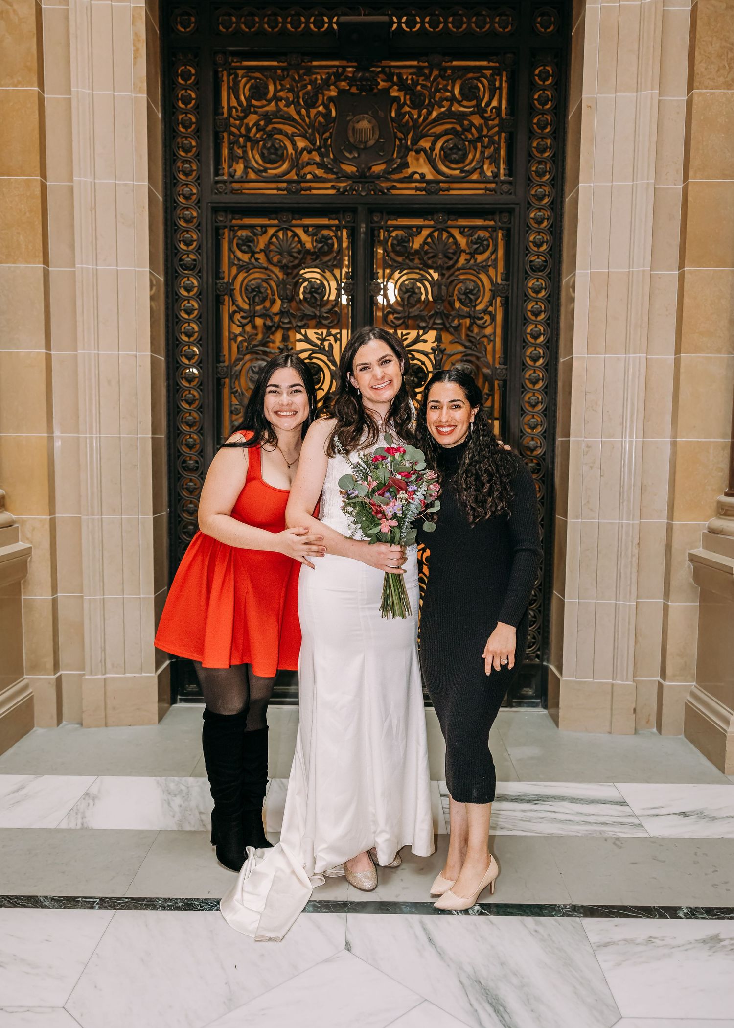 Bride in white gown holds bouquet flanked by two guests in red and black dresses before ornate golden doors.