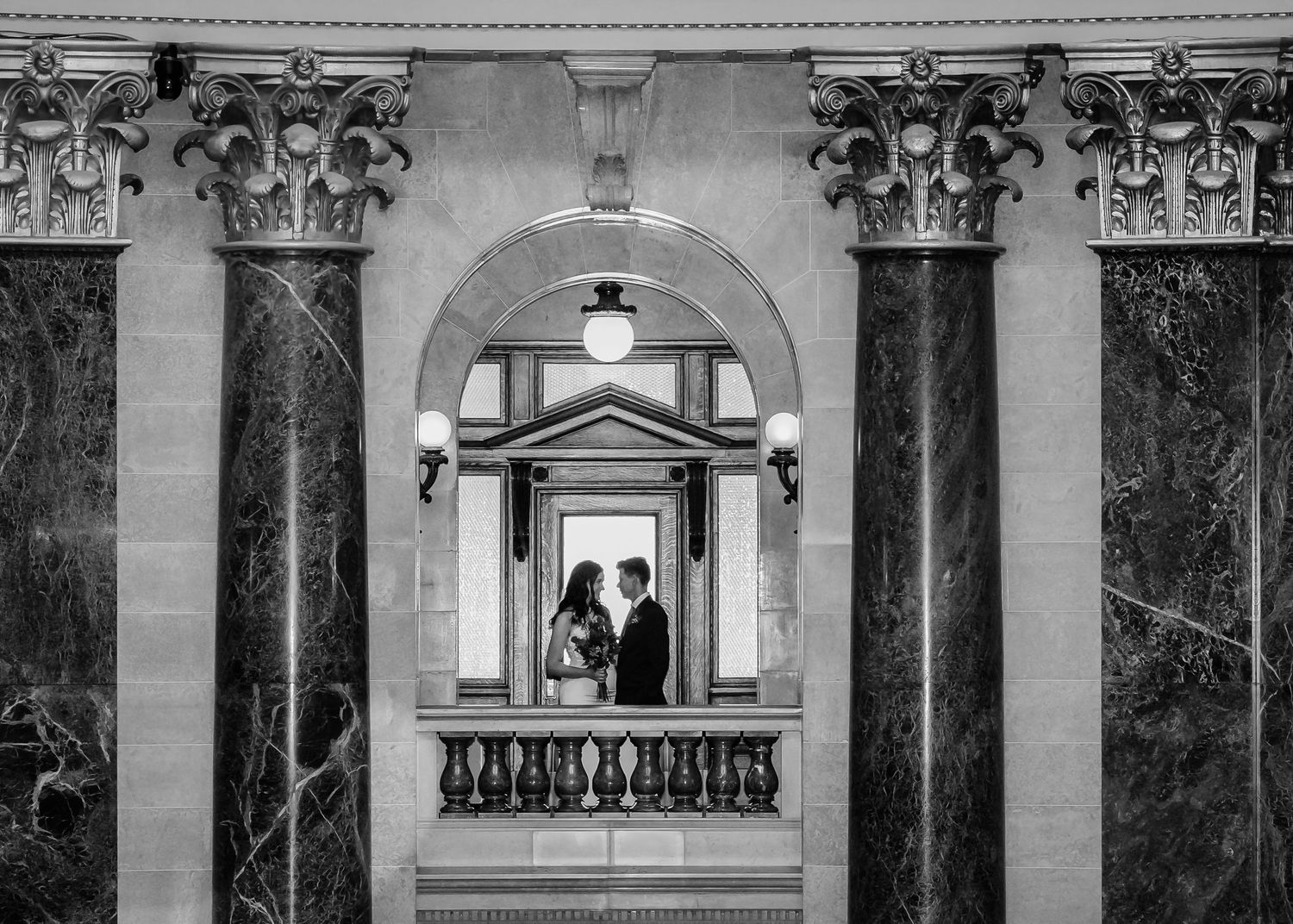 Black and white photo of couple framed by marble columns and arched doorway in elegant hall.