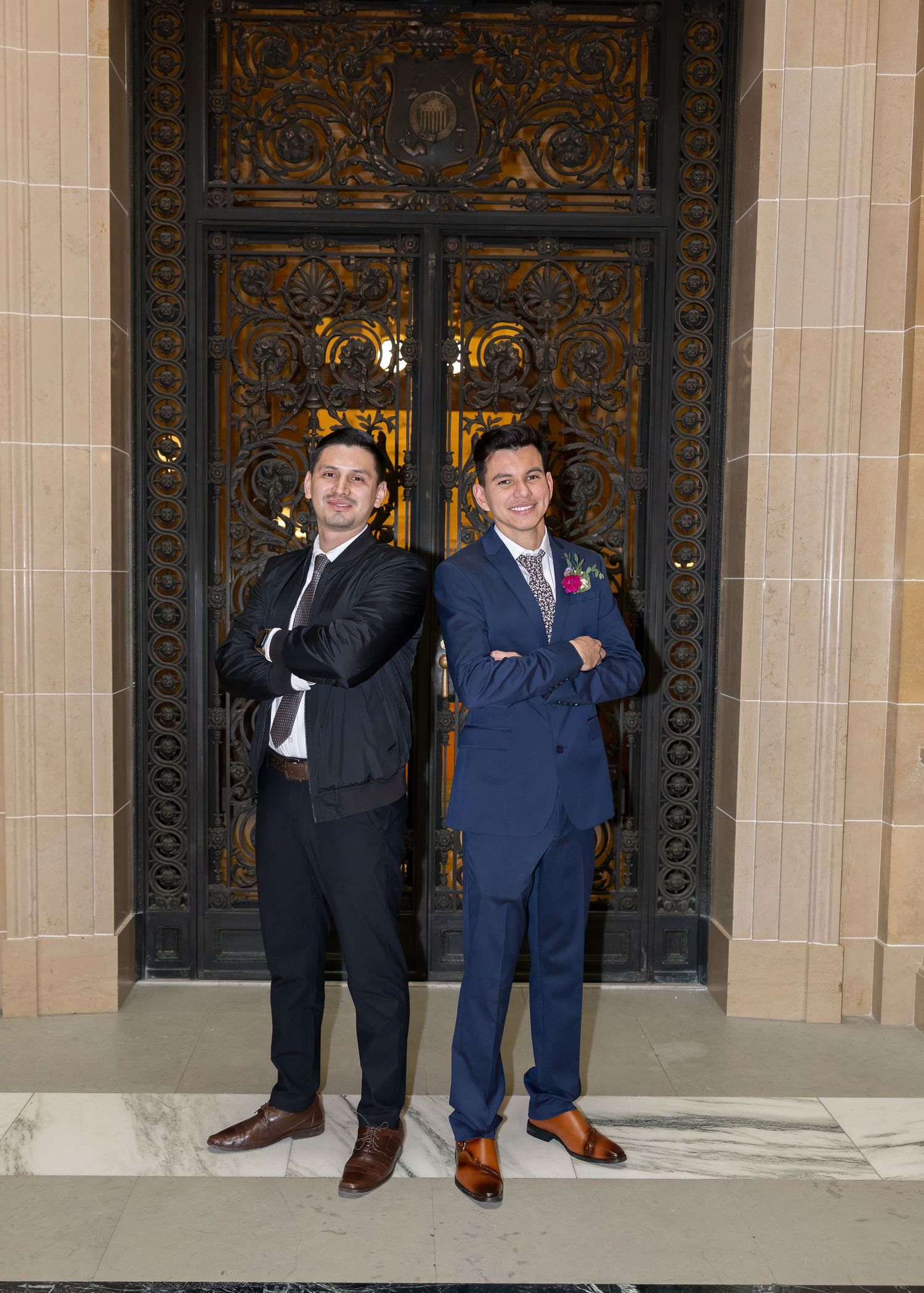 Two men in suits standing arms crossed before grand ornate bronze doors at a wedding venue.
