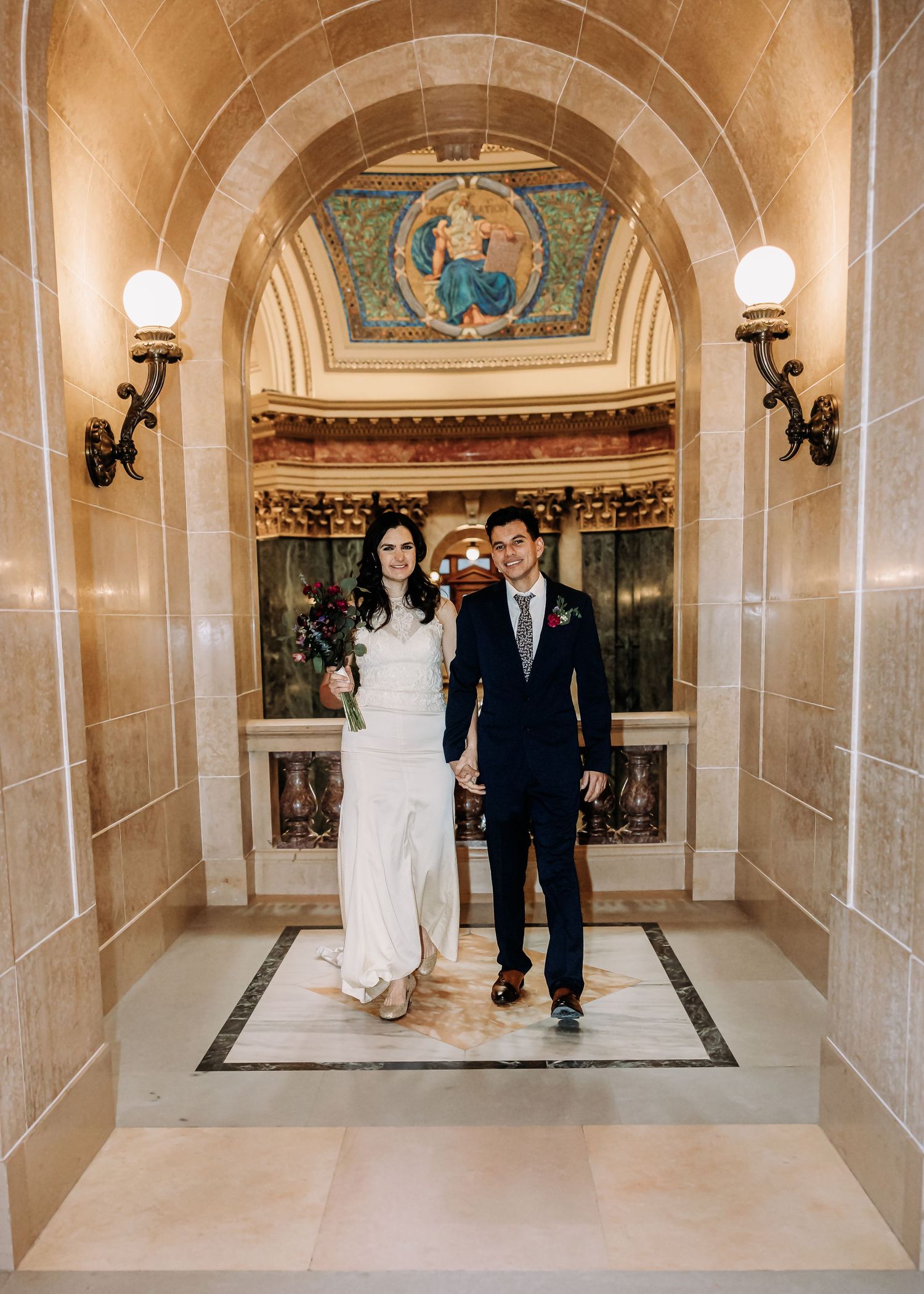 Bride holding bouquet and groom in navy suit walking hand in hand through elegant arched hallway of historic building.