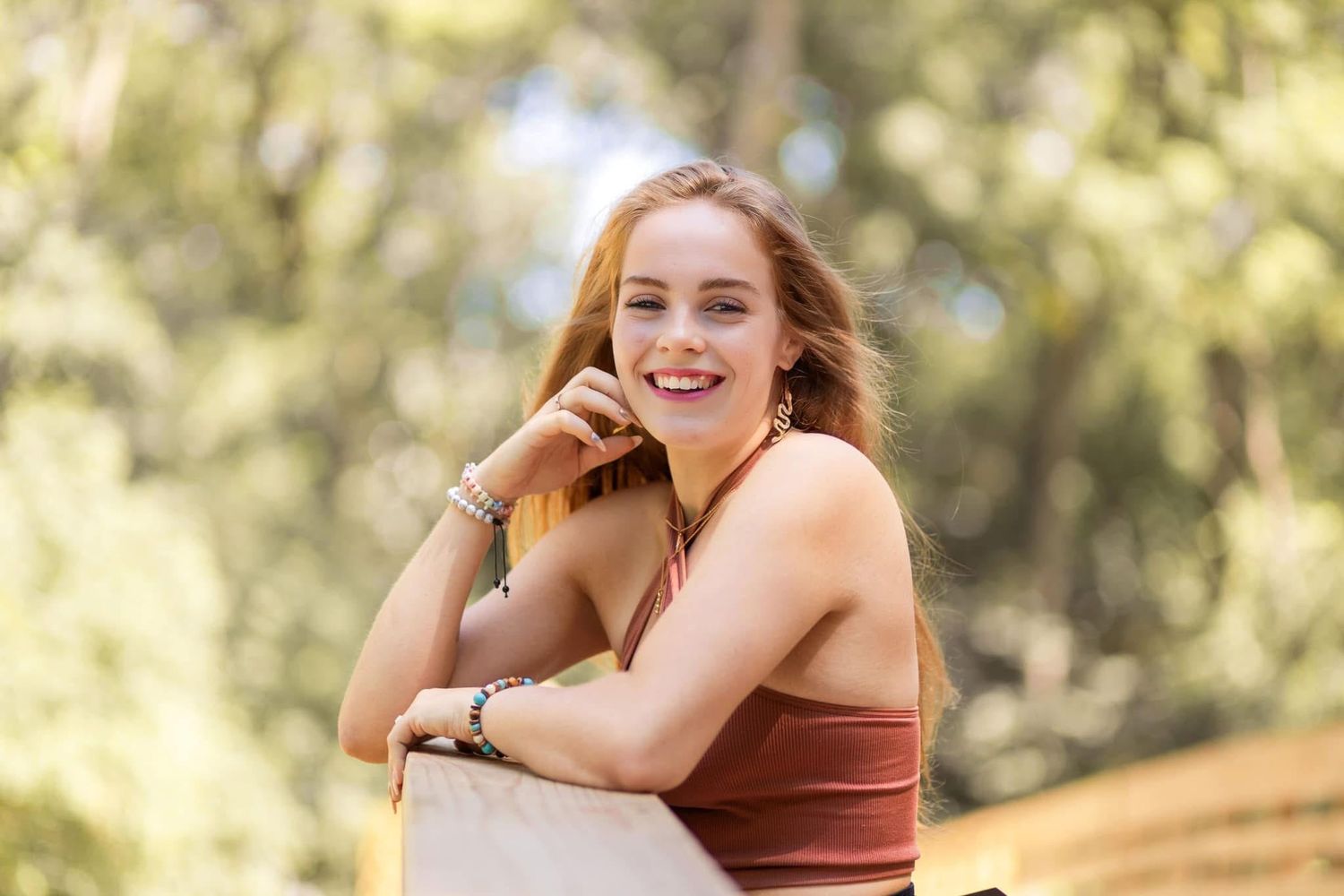 Smiling young woman with auburn hair leans on railing outdoors surrounded by sunlit green trees.