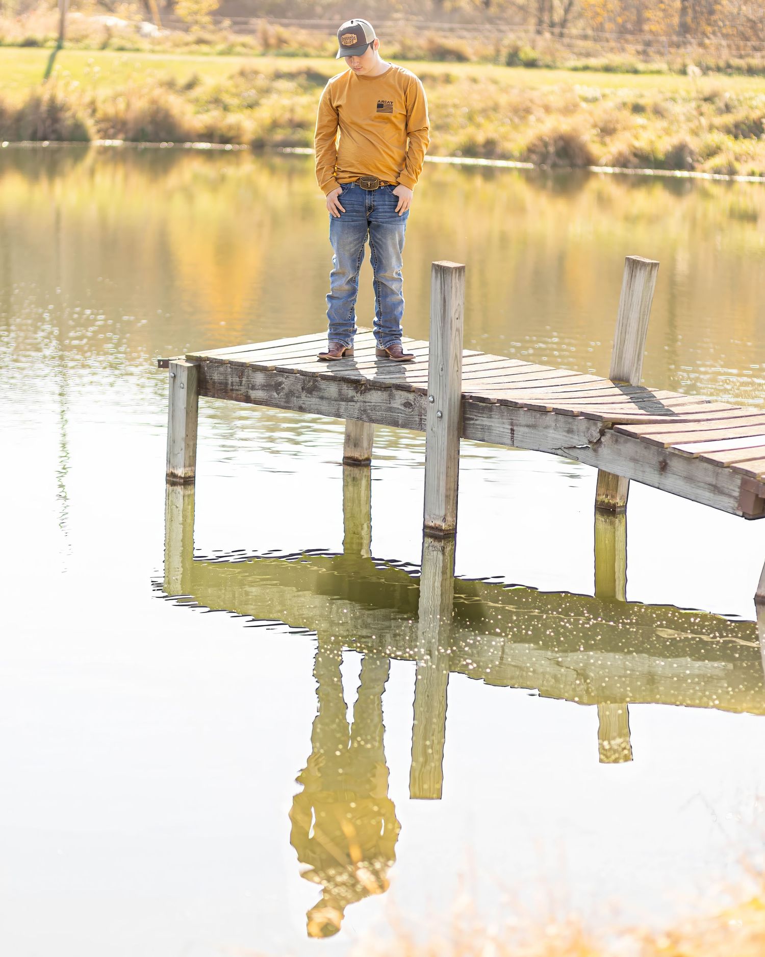 Senior boy in yellow shirt stands on wooden dock with reflection in calm lake.