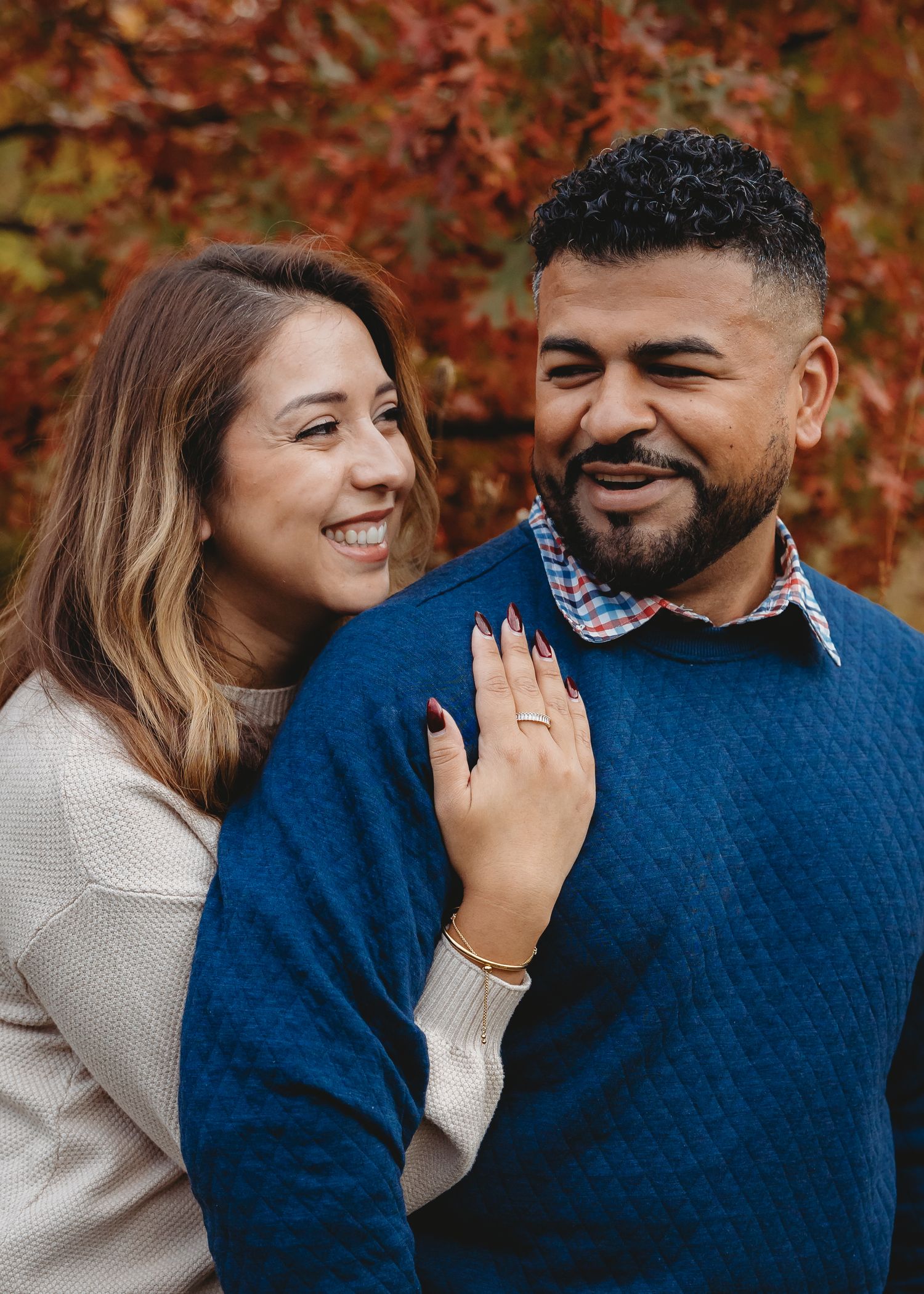 Couple smiling together in front of vibrant red autumn foliage during fall engagement photo session.