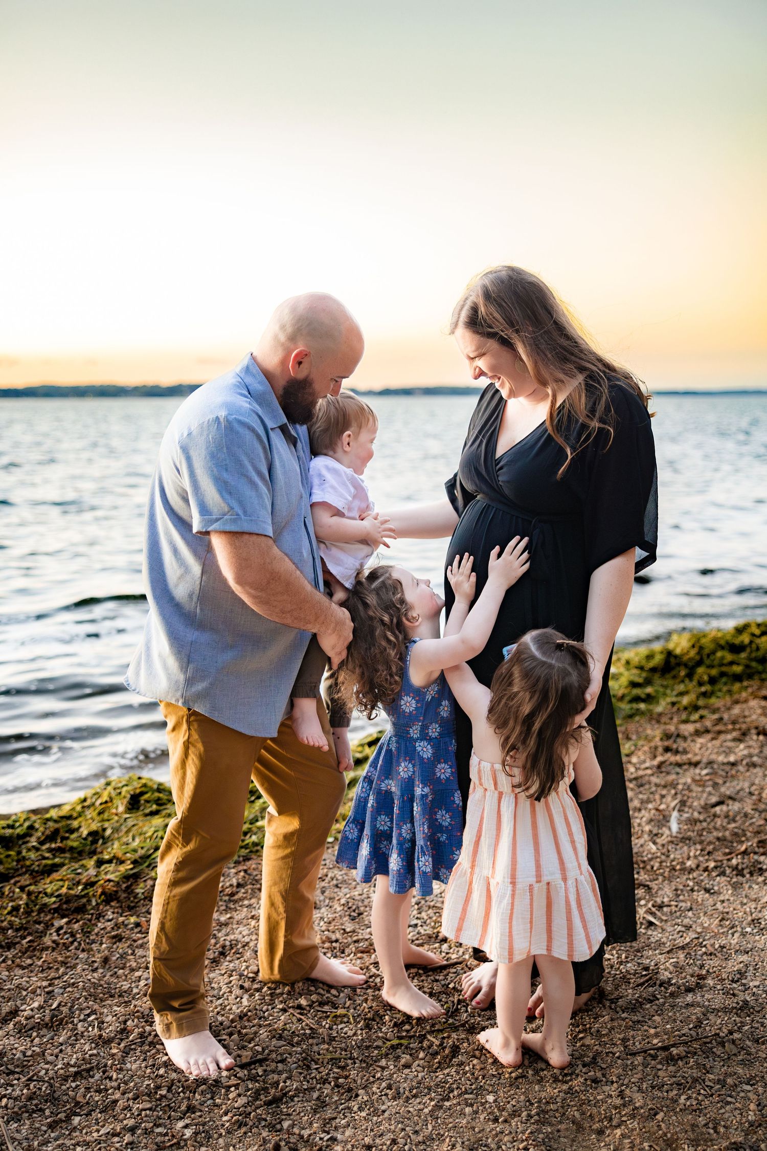 Pregnant woman in black dress with family on beach at sunset, husband holding baby while two girls touch her belly.