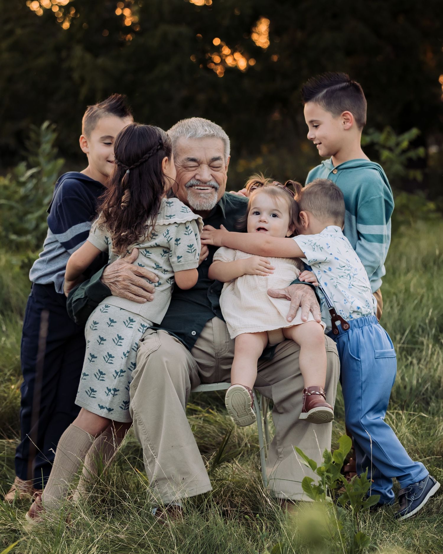 Grandfather joyfully embraced by five grandchildren outdoors in a lush green field during golden hour.