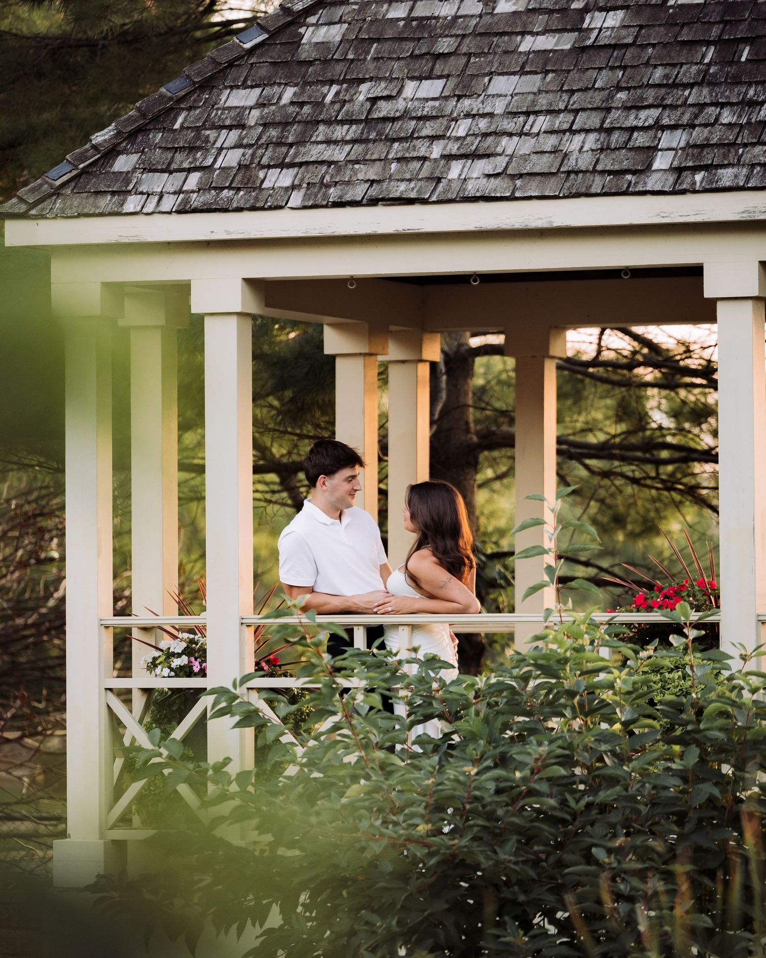 A couple shares a romantic moment inside a charming white gazebo surrounded by lush greenery and red flowers.