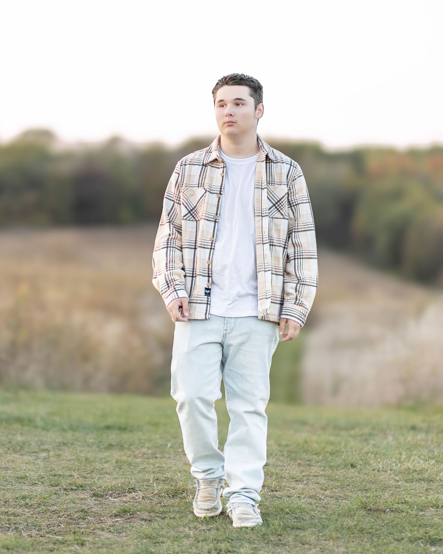 Senior boy in plaid flannel shirt walks through green field during outdoor photo session.