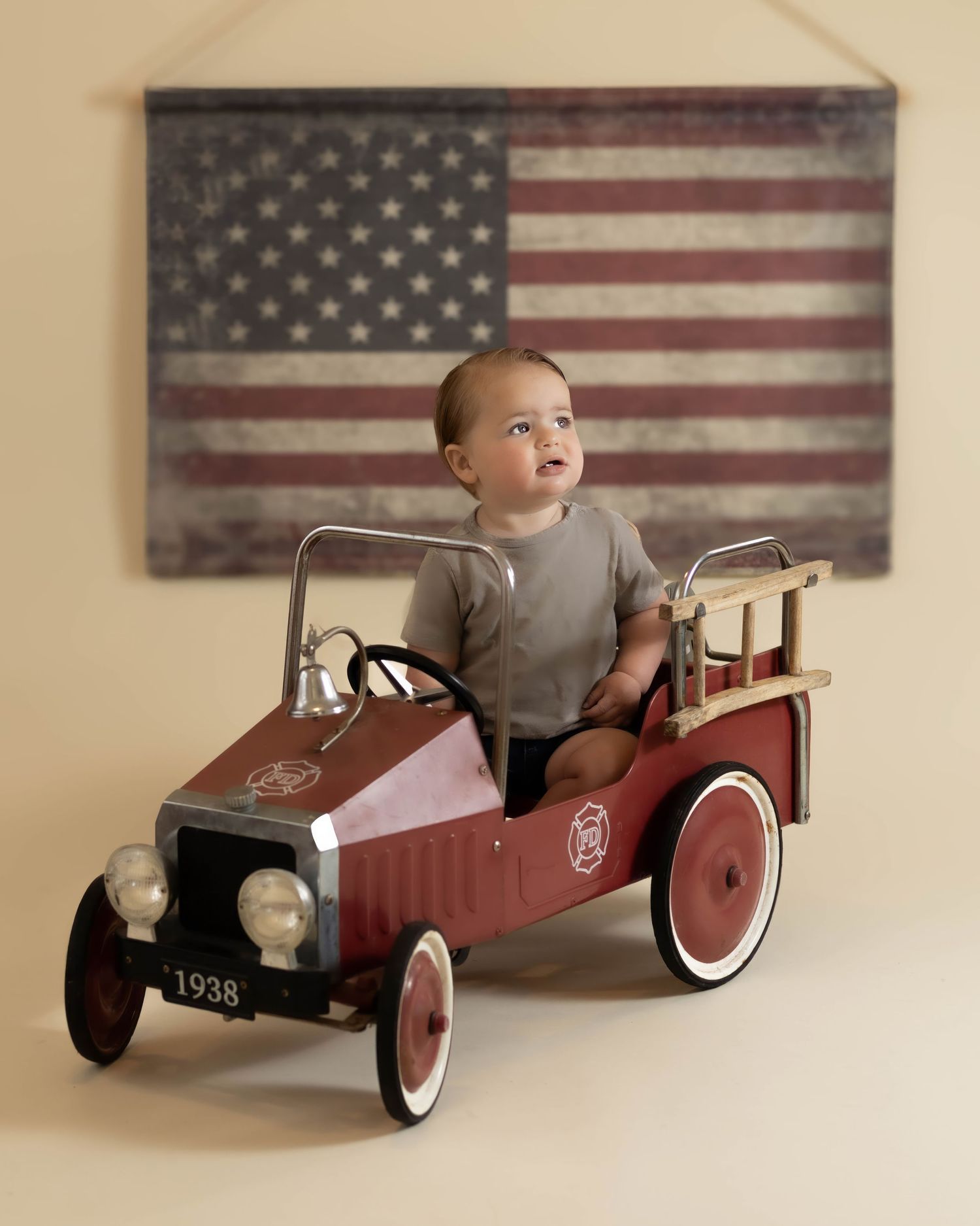 Baby boy in gray onesie sits in a vintage 1938 red pedal fire truck before an American flag.