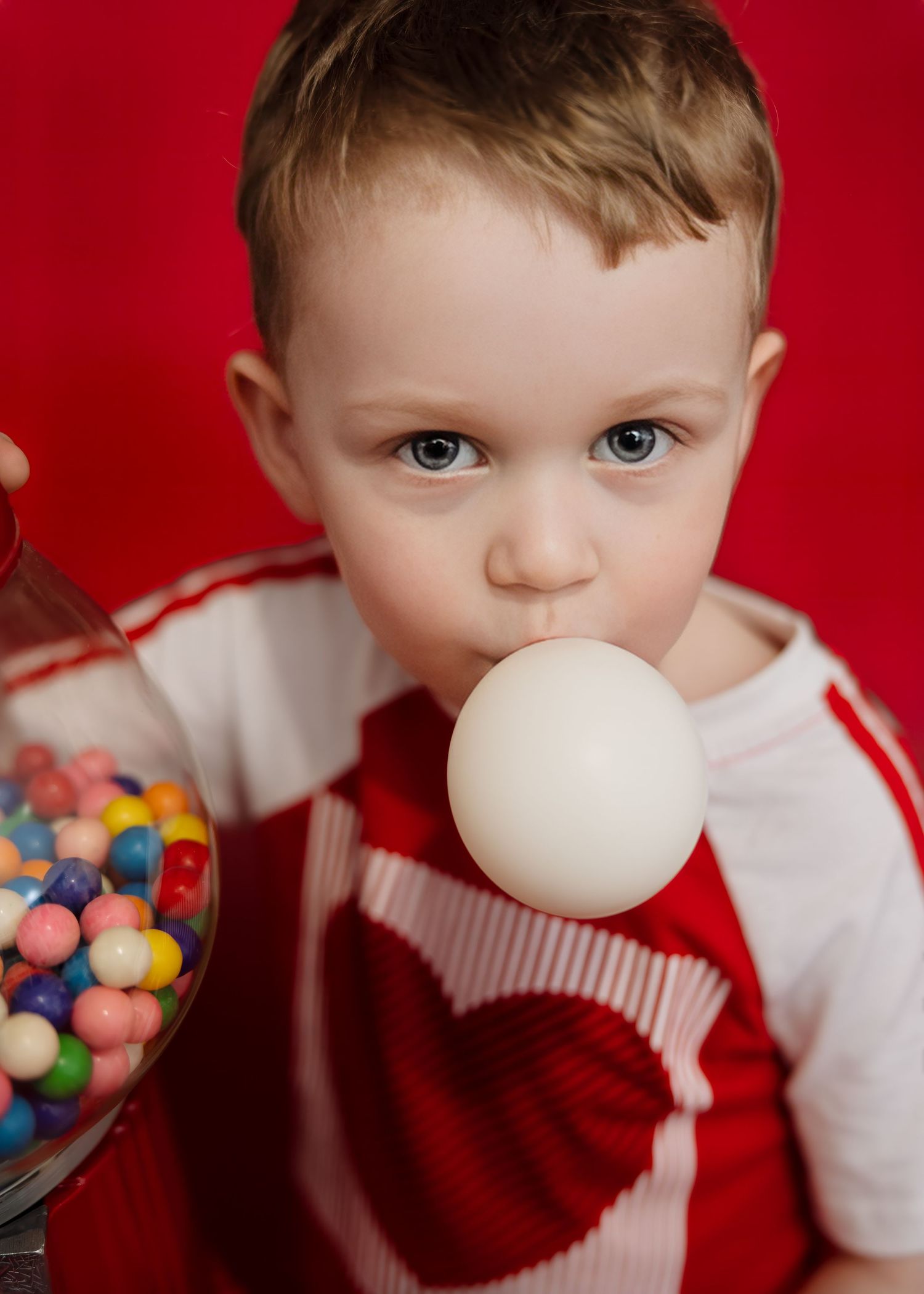 Toddler boy blows bubble gum bubble near colorful gumball machine on red background.