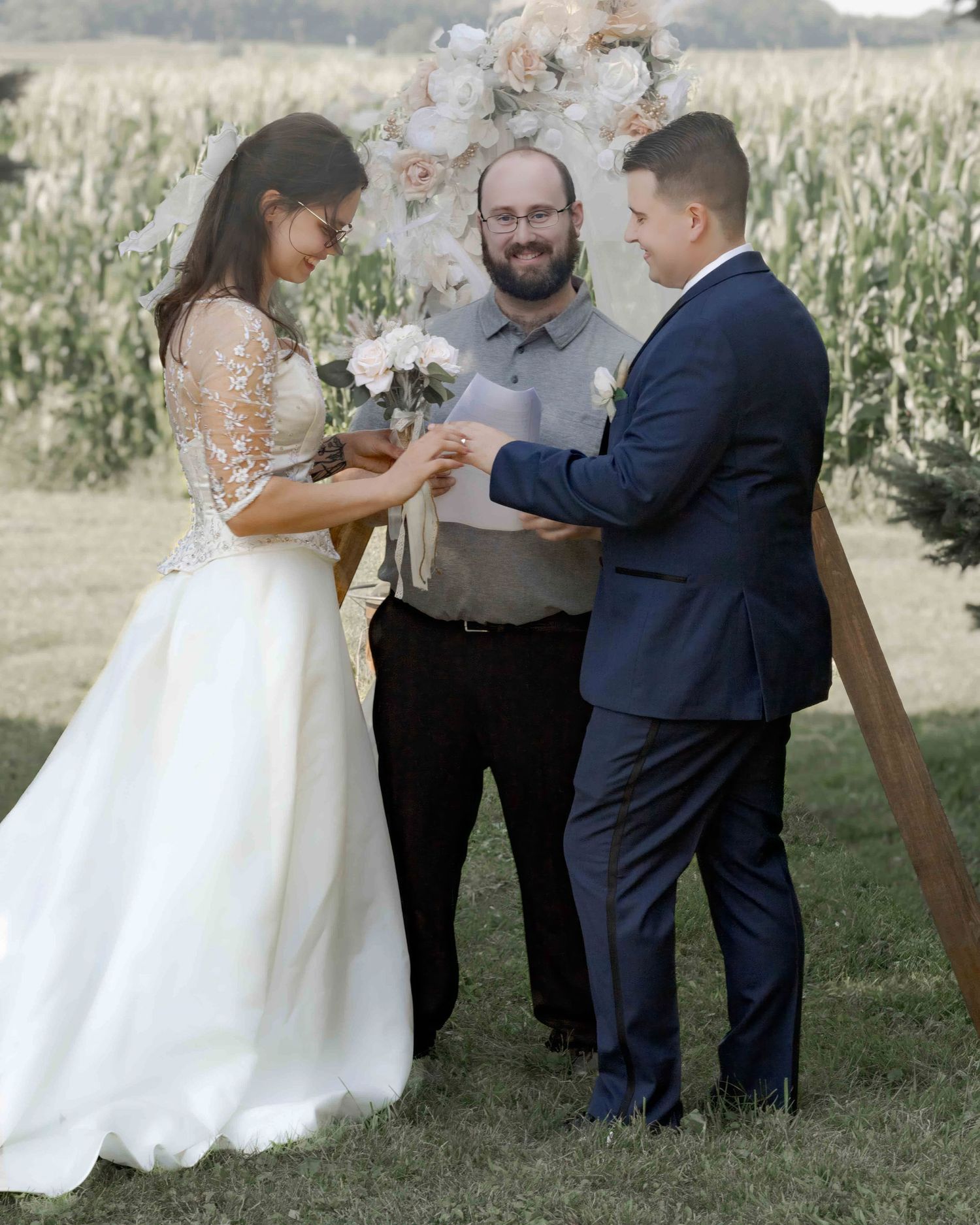 Bride places ring on groom's finger during outdoor wedding ceremony near cornfield.