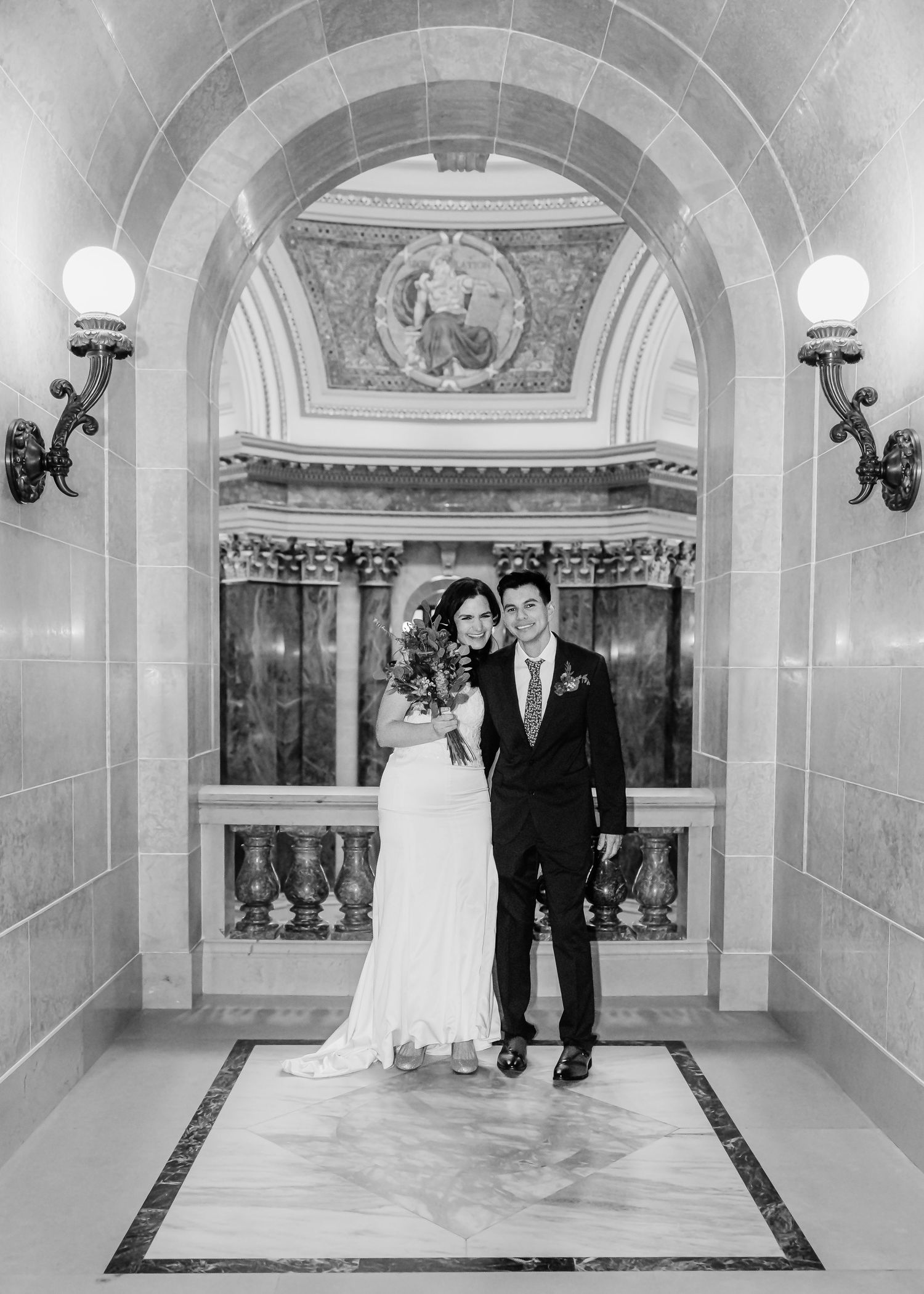 Black and white formal wedding portrait of bride and groom standing under decorative stone arch with mosaic detail.