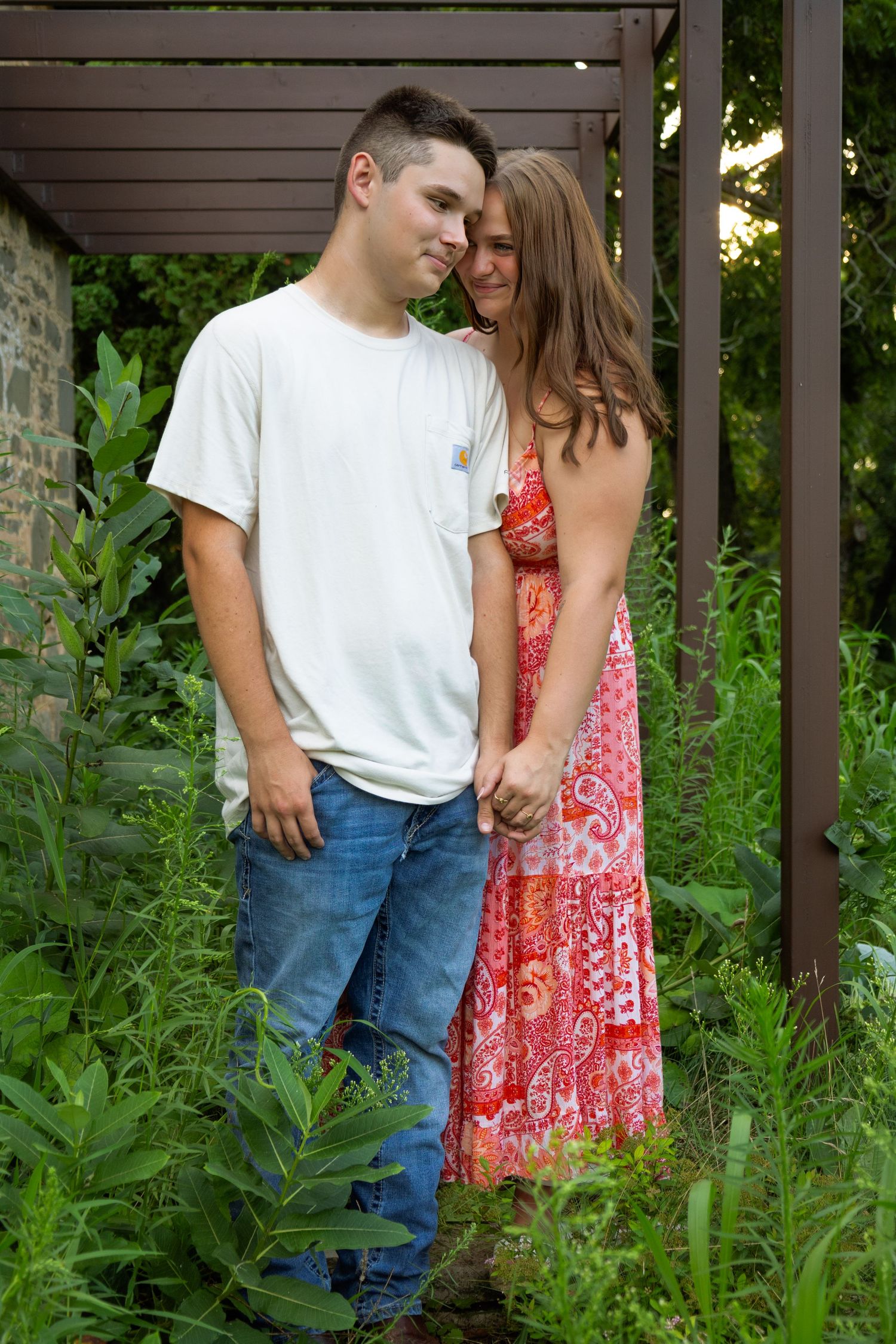 Young couple standing close together holding hands in a lush green garden under a pergola.