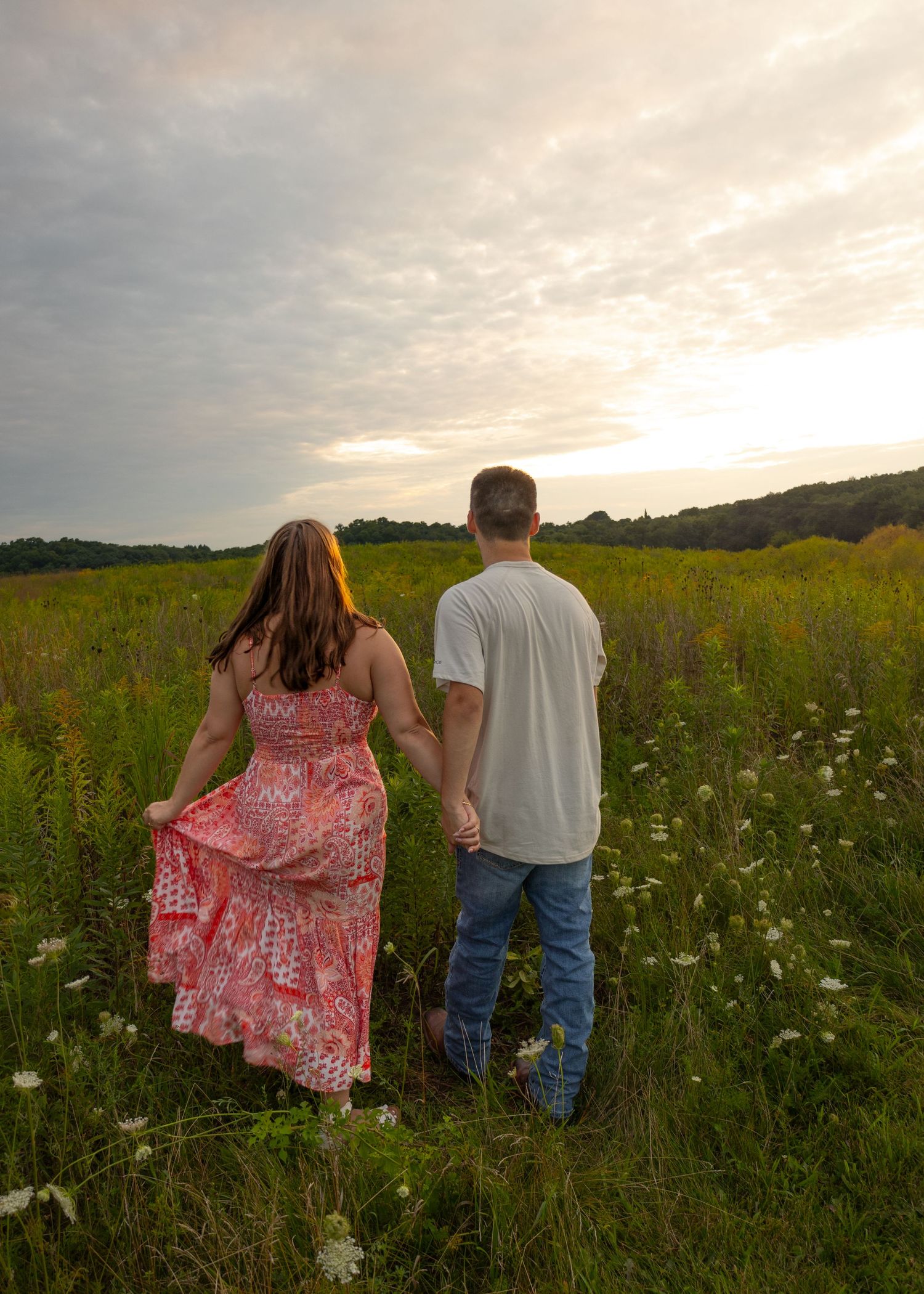 Couple holding hands walking through a wildflower meadow at sunset, woman in coral maxi dress, man in white tee.