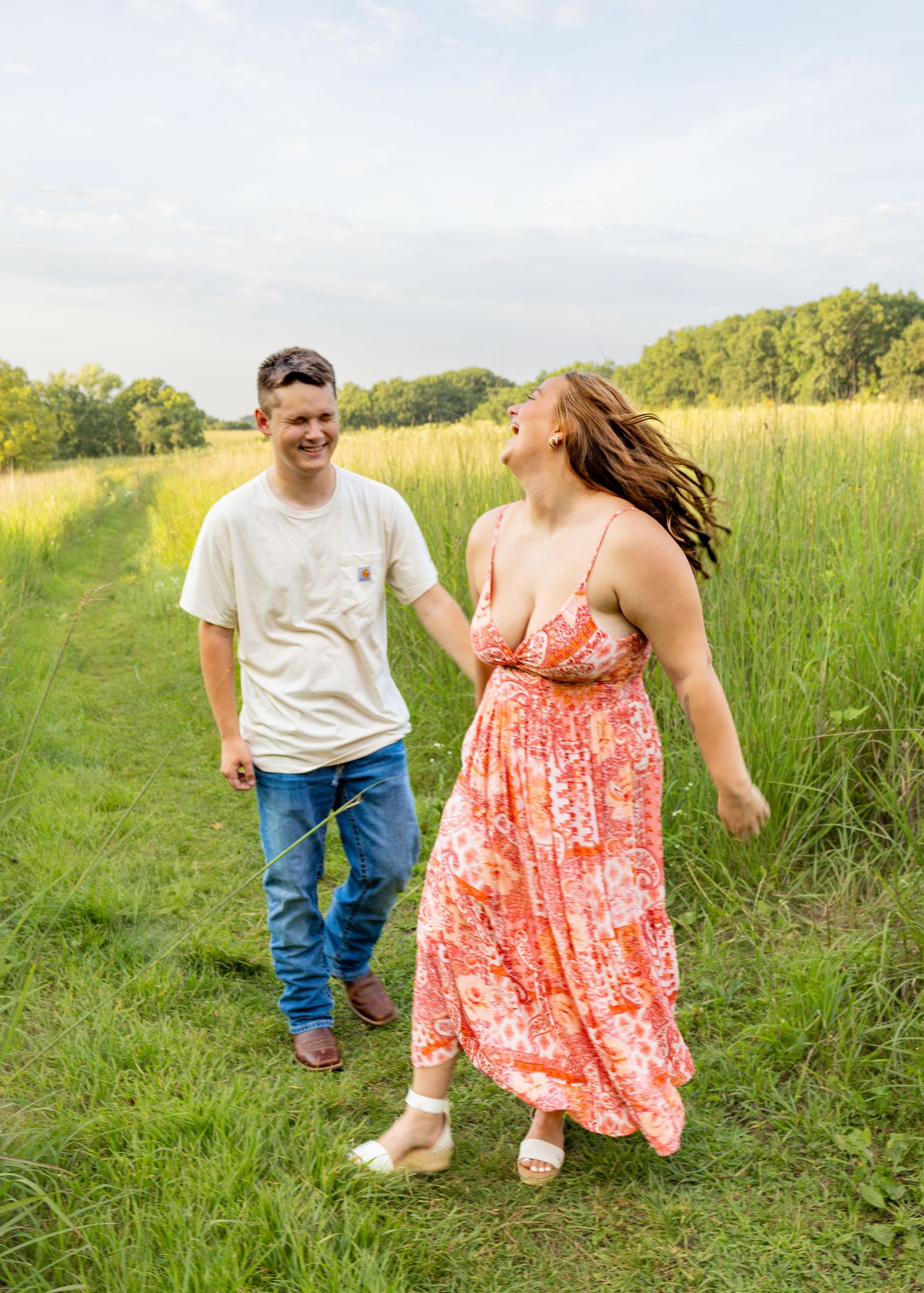 Joyful couple laughing and walking hand in hand through lush green meadow path on a sunny day.