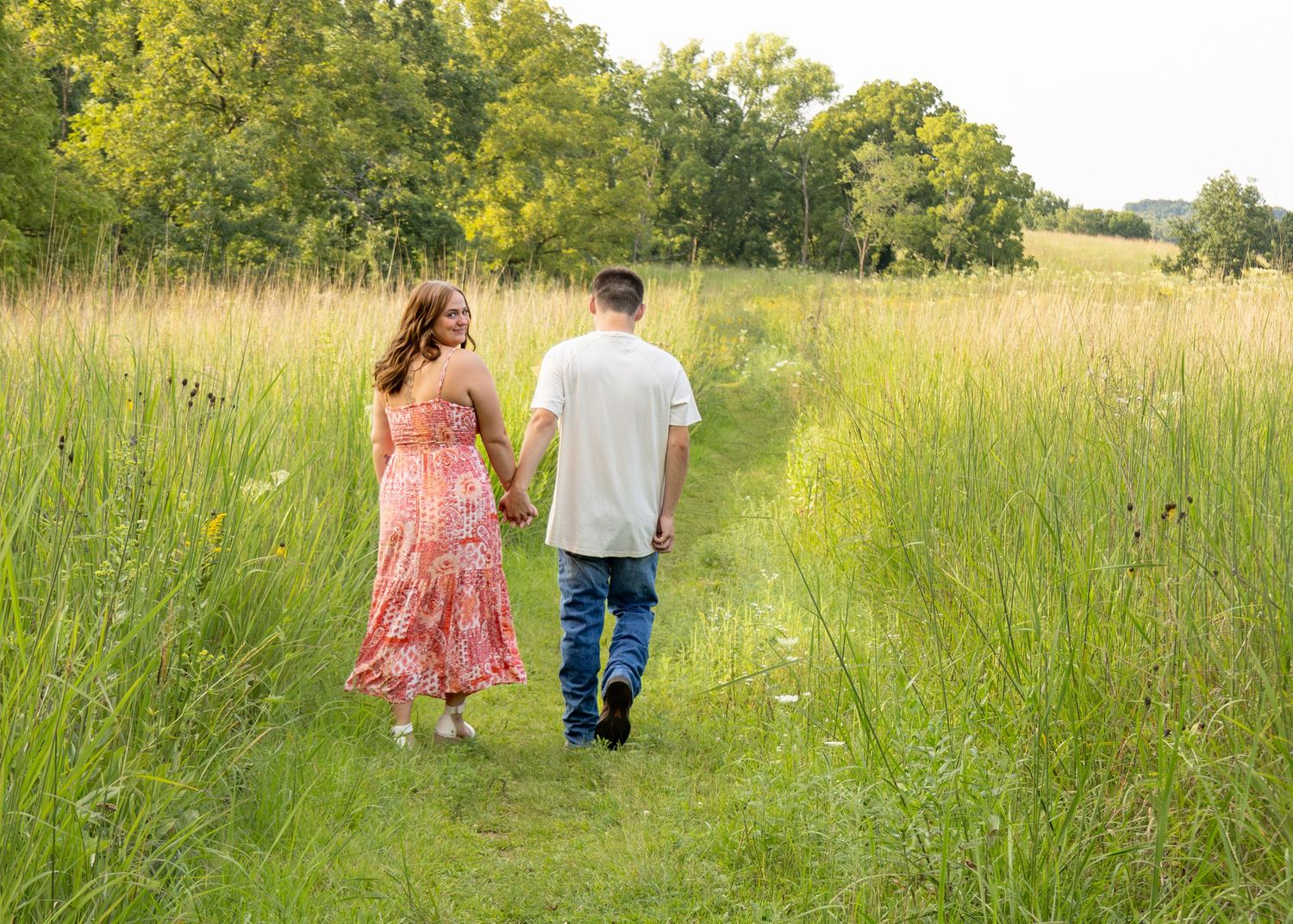 A romantic couple walks hand-in-hand away from the camera along a scenic green meadow trail.