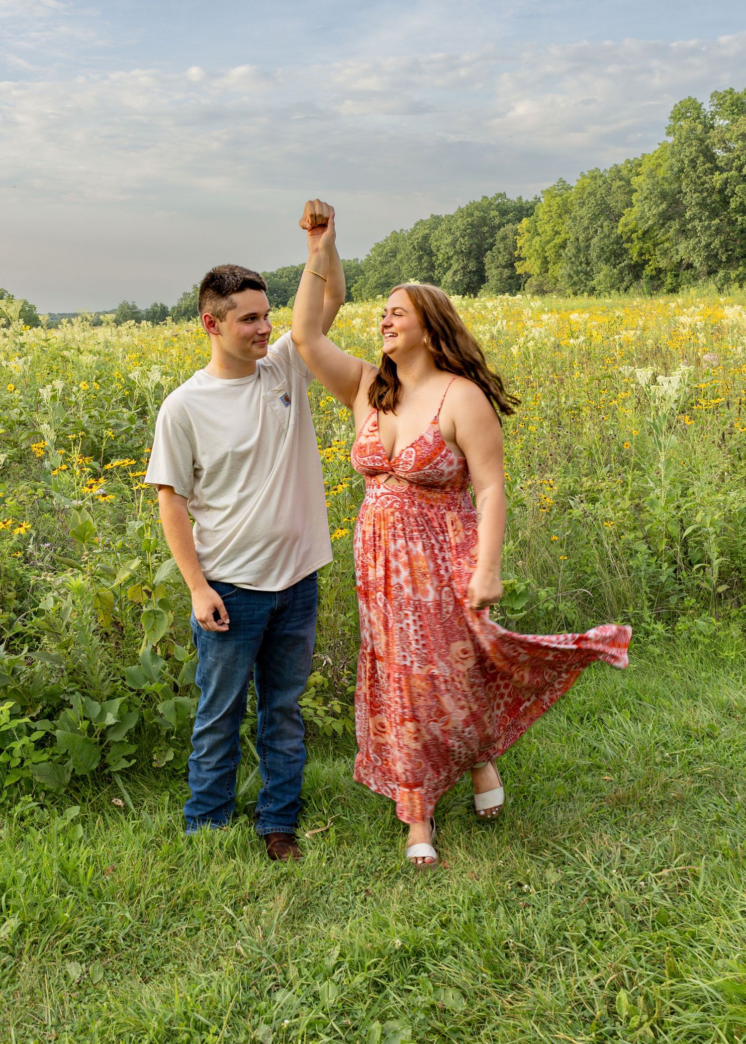 Smiling couple dancing in a green meadow with yellow wildflowers under a cloudy sky.
