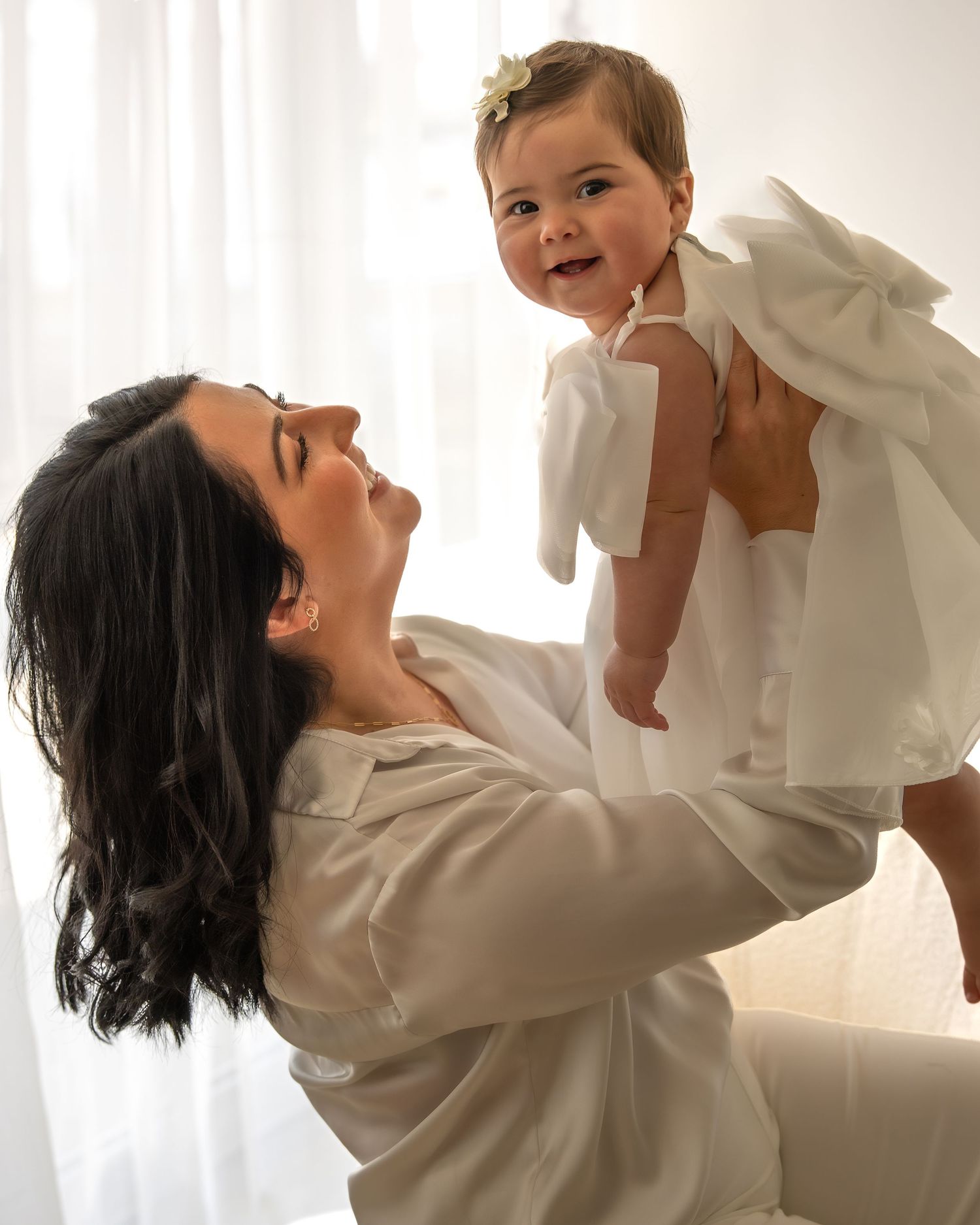 Joyful dark-haired mother lifting smiling baby girl in white bow dress in bright white room.