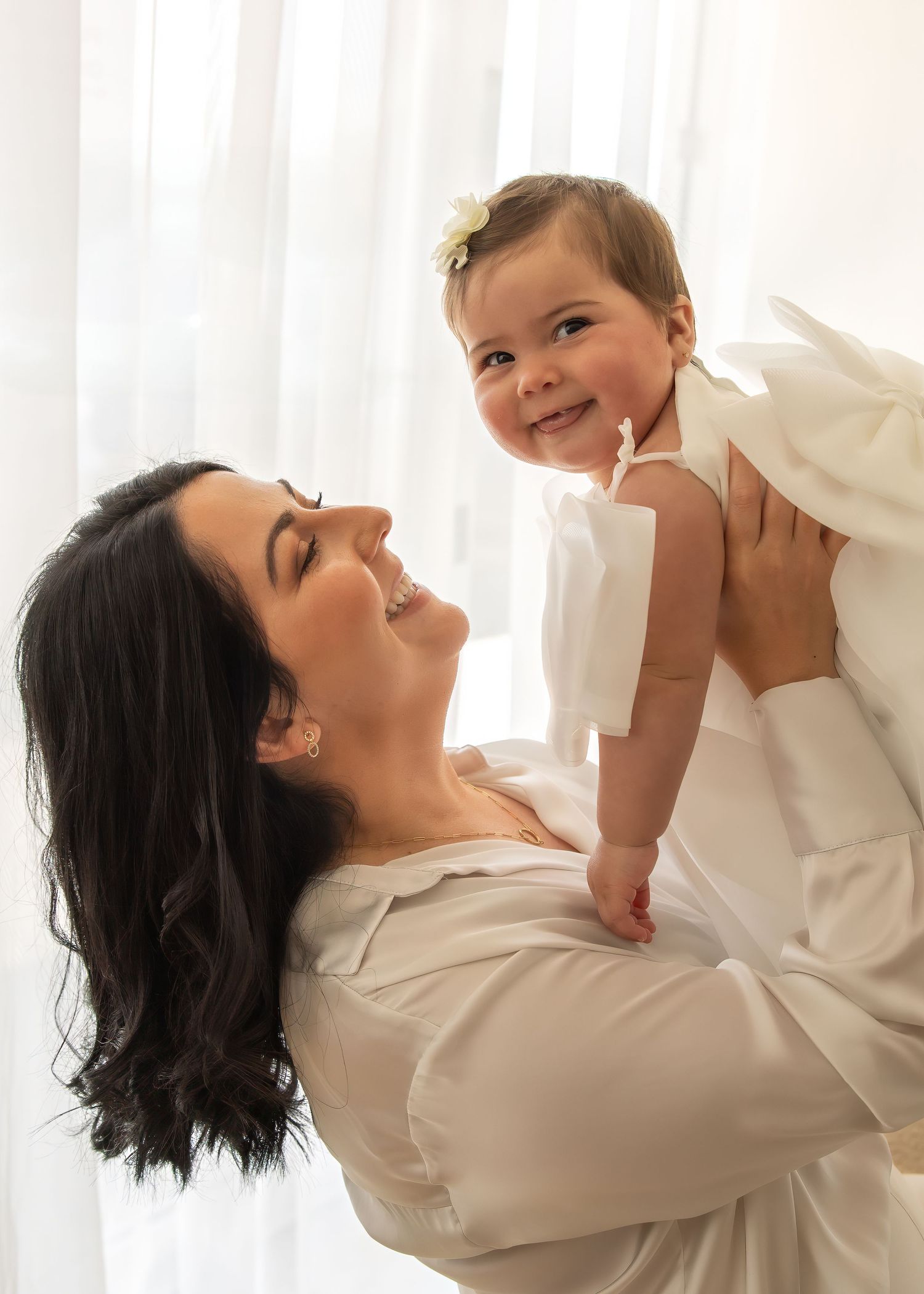 Joyful mother in white lifting smiling baby girl in white dress near bright window indoors.