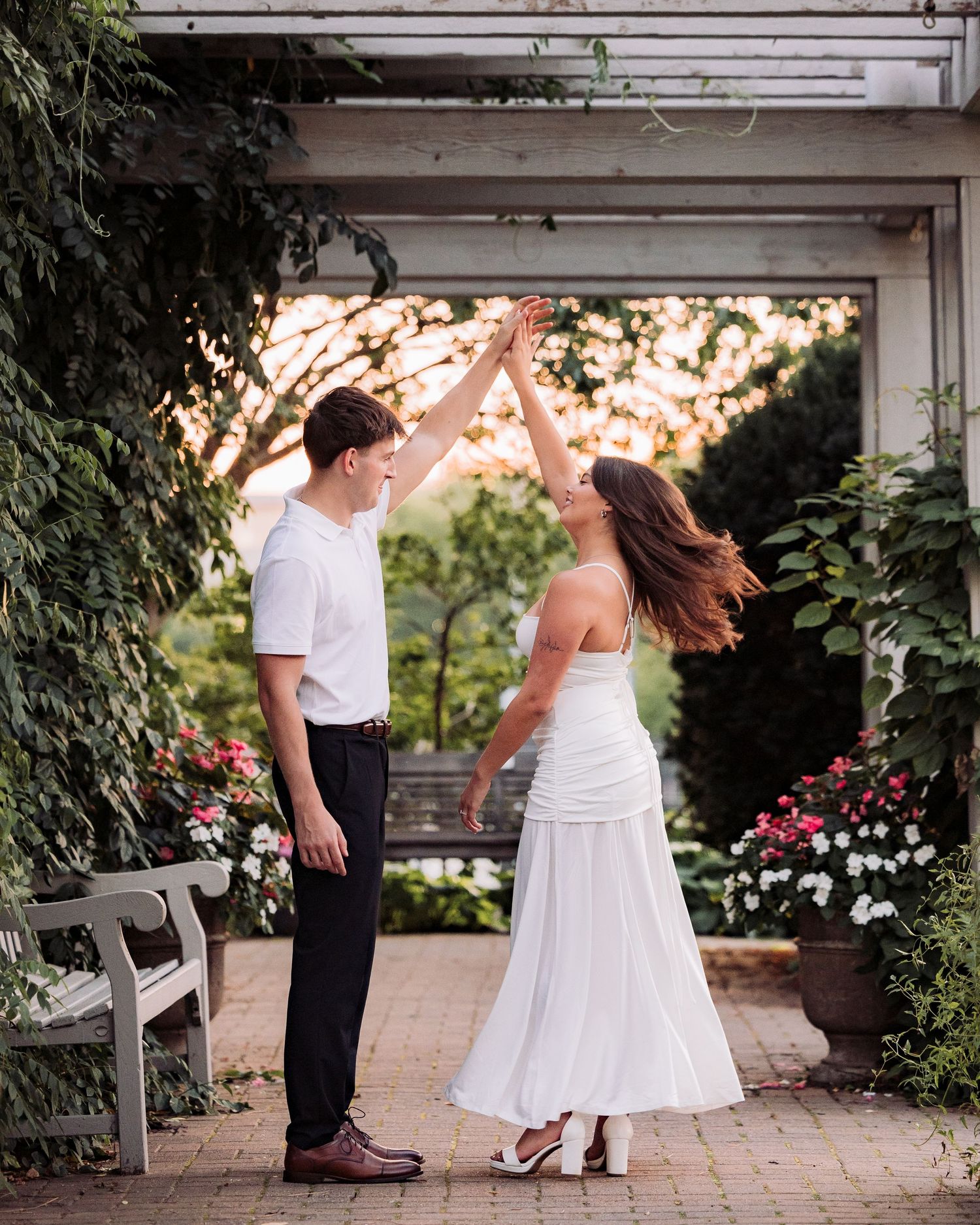 Couple dancing outdoors under a garden pergola, man in white shirt spinning woman in white dress at golden hour.