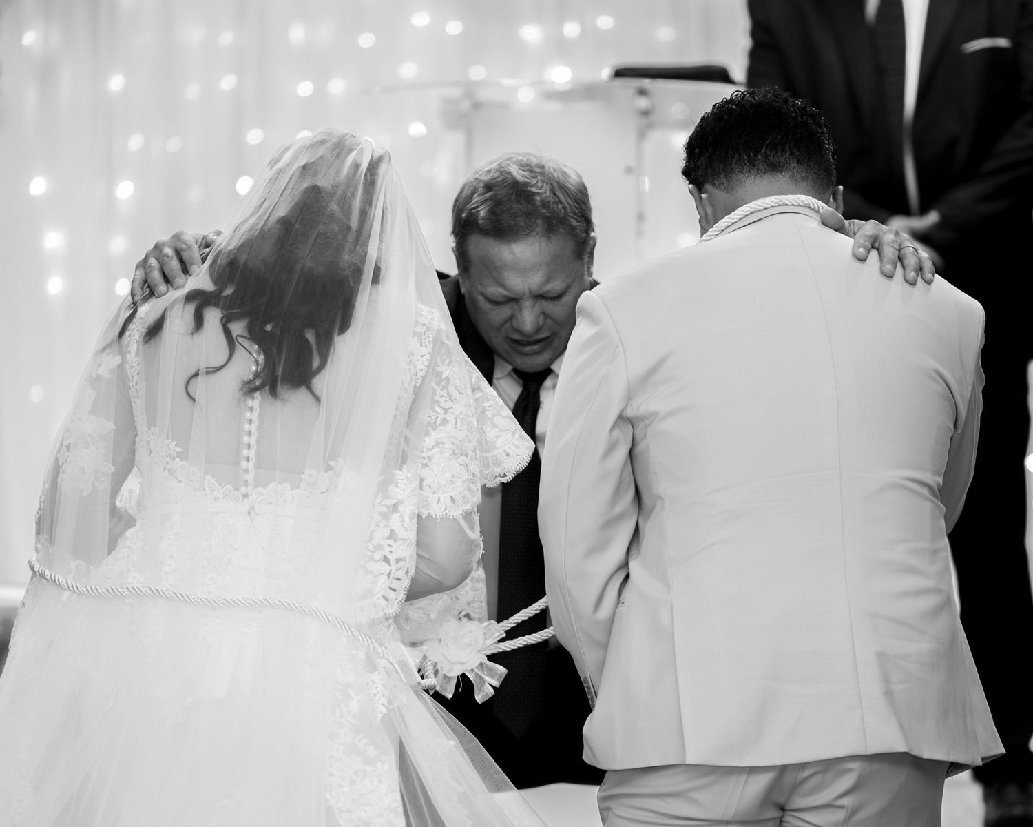Bride and groom kneel in prayer at a wedding ceremony while an officiant places his hands on their shoulders in a black and white photo.