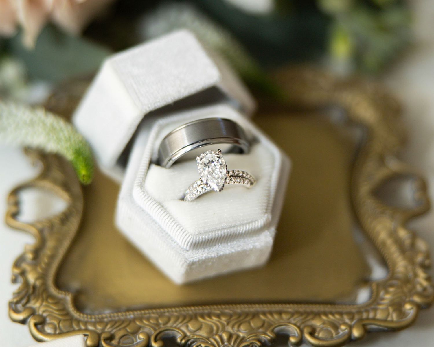 Wedding rings in a white velvet hexagonal box, featuring a diamond engagement ring and a men's band, on an ornate gold vintage tray.