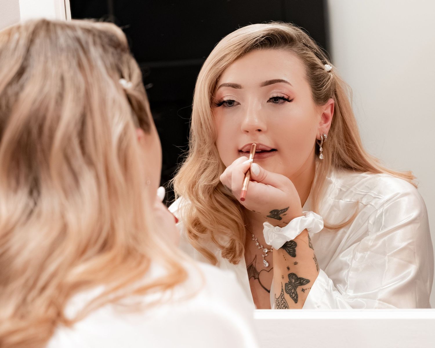 Blonde woman in a white satin robe applies lip makeup while looking into a mirror, showing arm tattoos and a white scrunchie on her wrist.