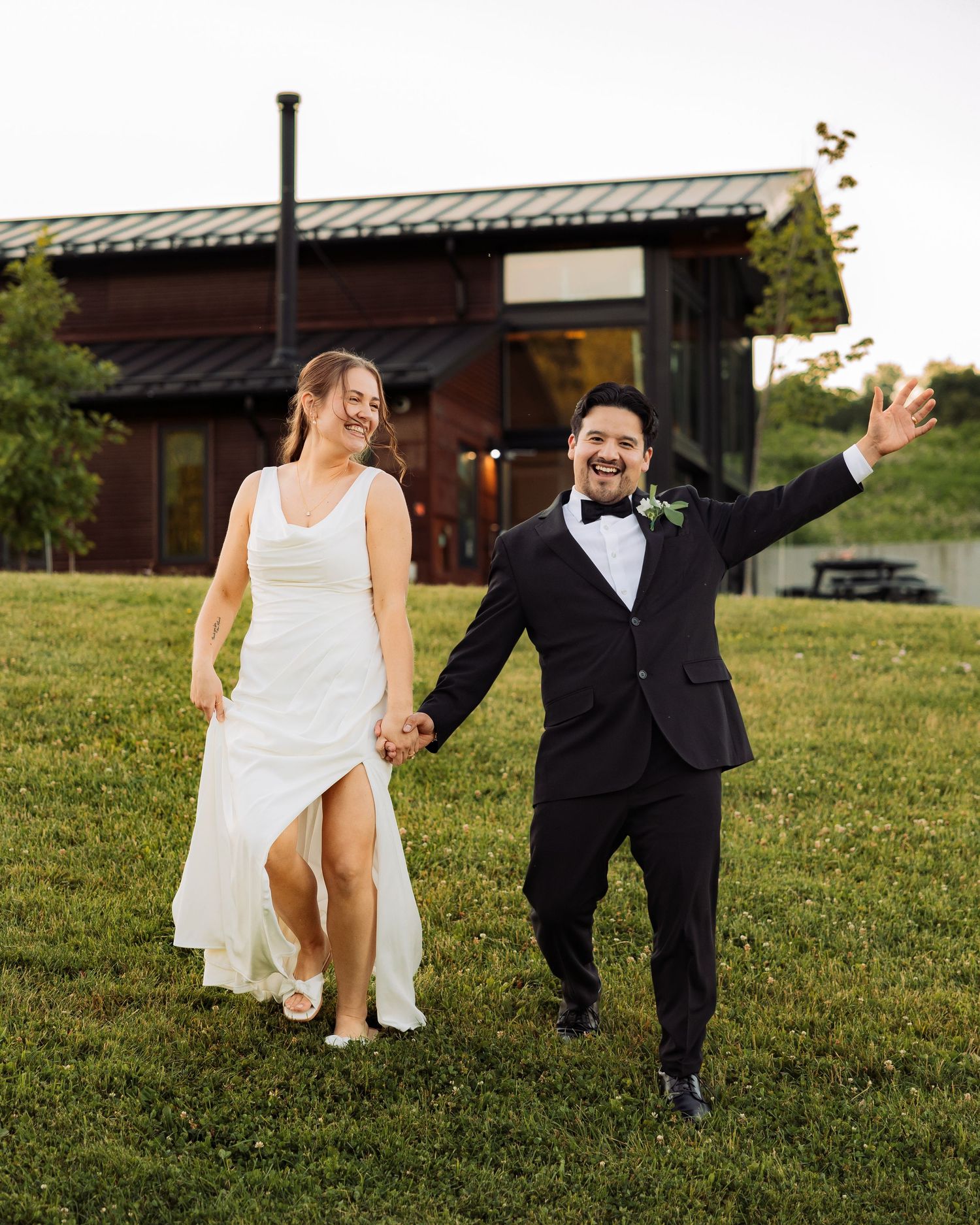 Happy newlyweds walk hand-in-hand on green lawn, bride in white gown, groom in black tuxedo, celebrating outdoors.