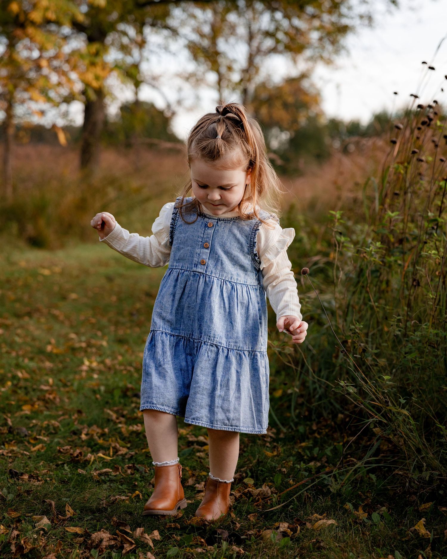 A toddler girl in a denim dress and brown boots plays outdoors among autumn leaves and tall grass.