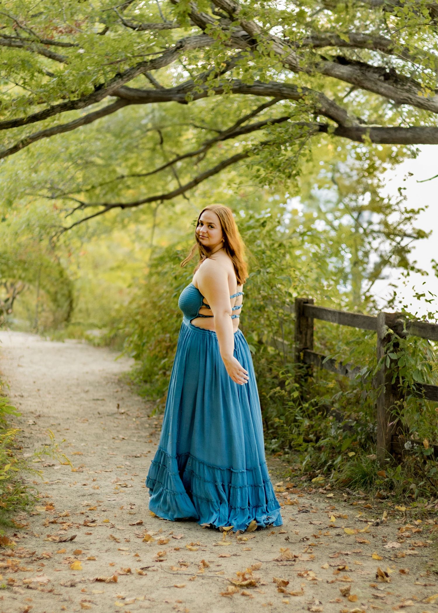 Senior girl in flowing teal gown stands on tree-lined path turning back for elegant portrait.