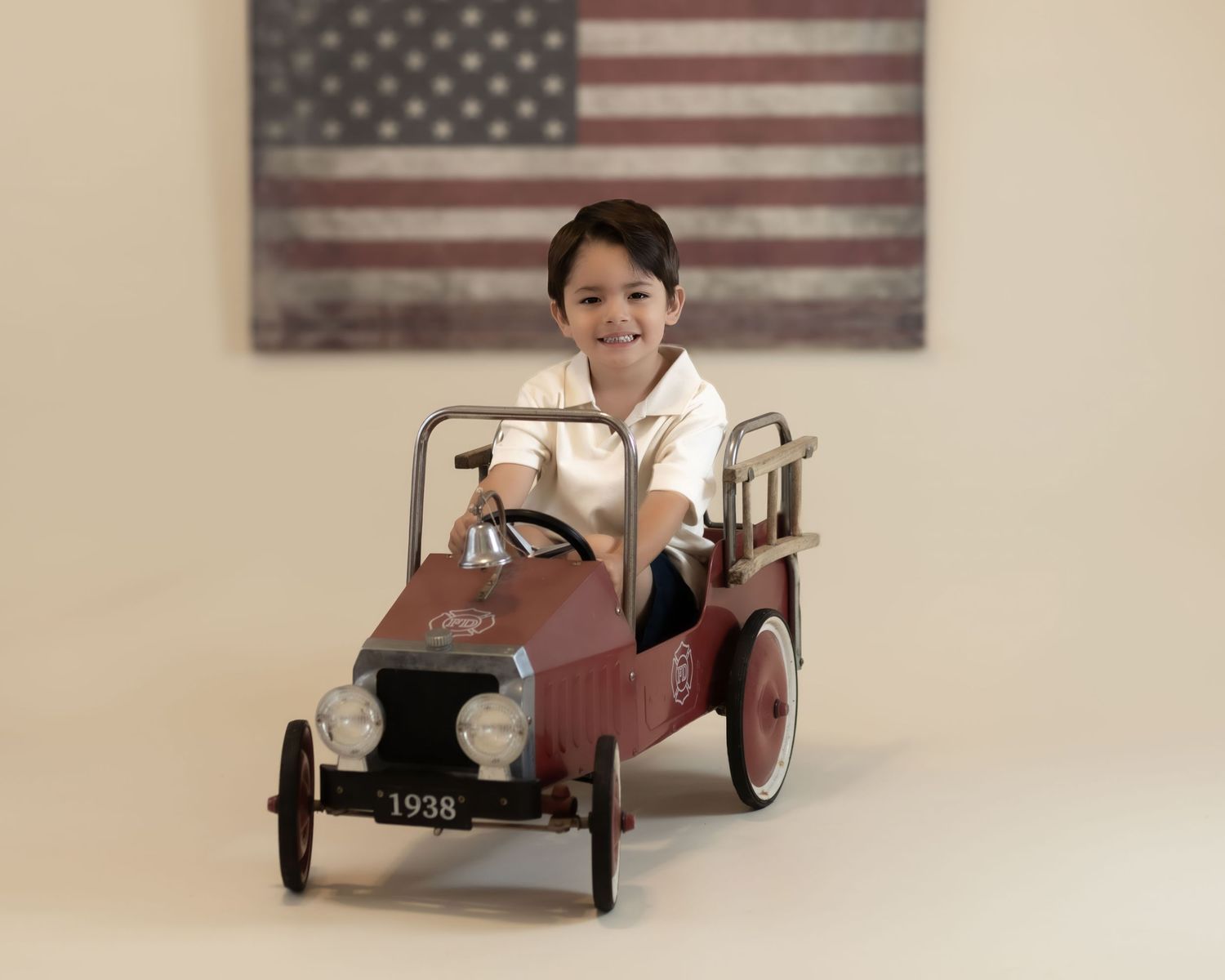 Young boy in white shirt smiles while riding a vintage 1938 red pedal fire truck before an American flag.