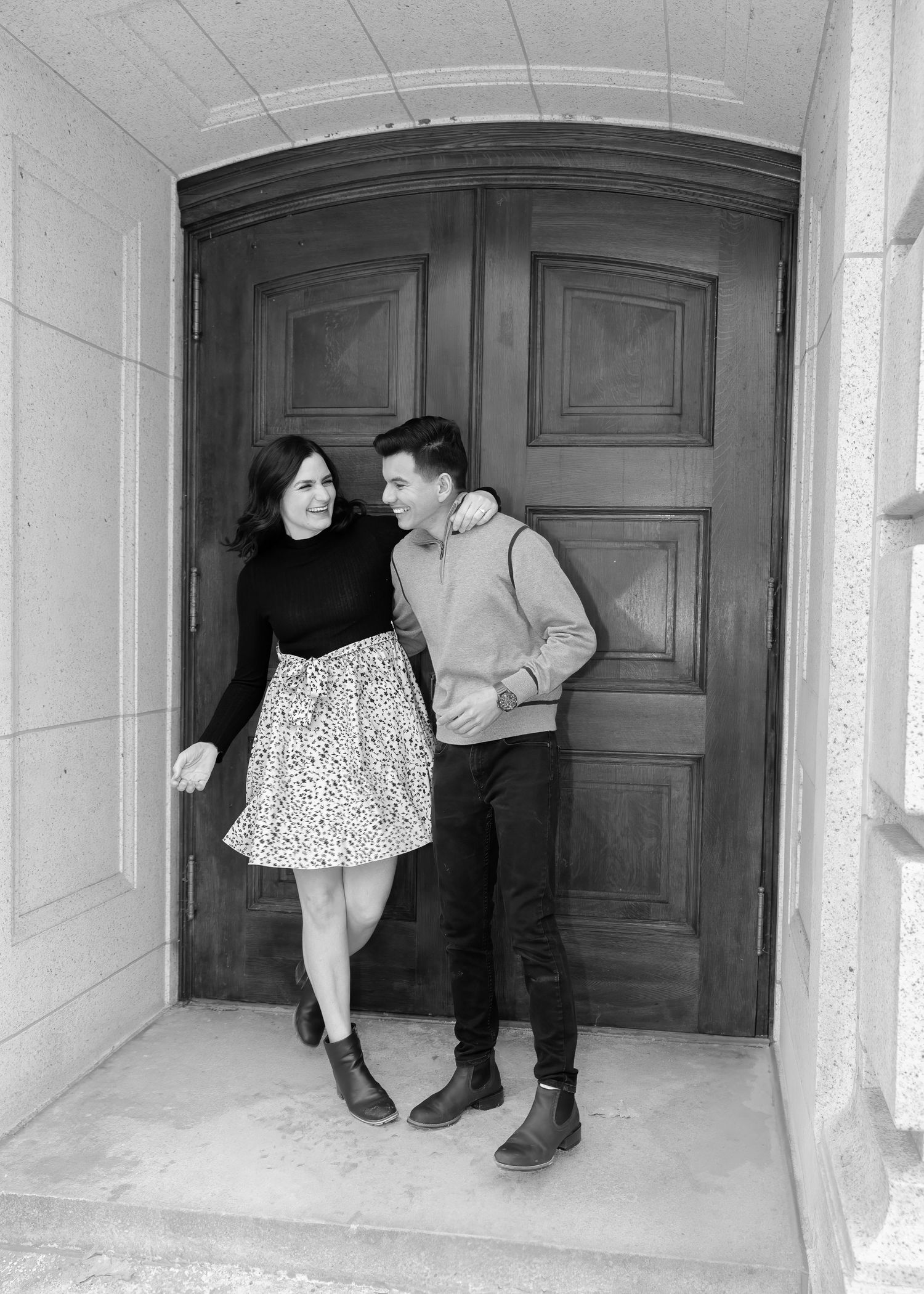 Black and white photo of a smiling couple leaning against large wooden double doors looking at each other.