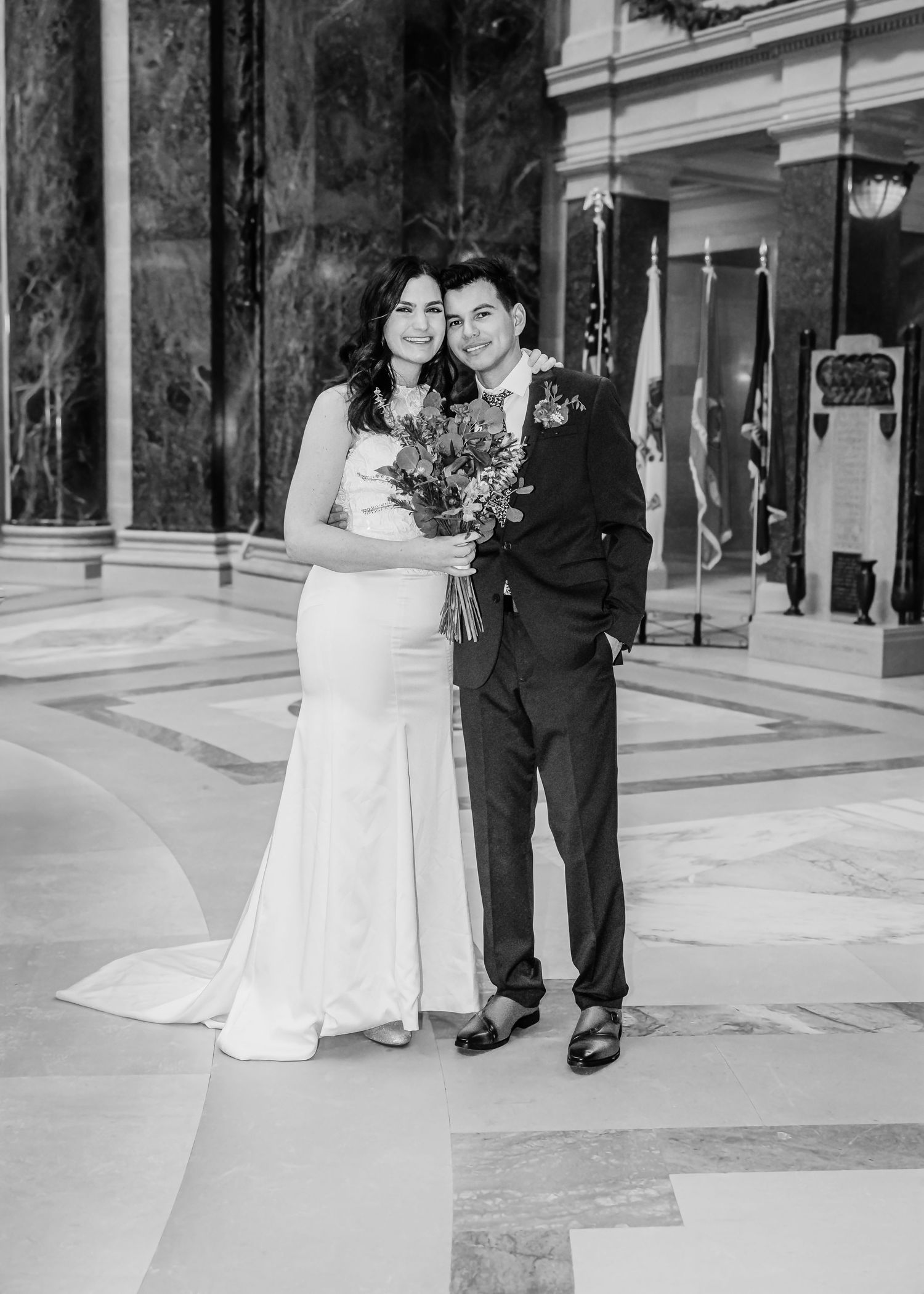Newlywed couple poses in marble hall, bride in white gown holding bouquet beside groom in dark suit.