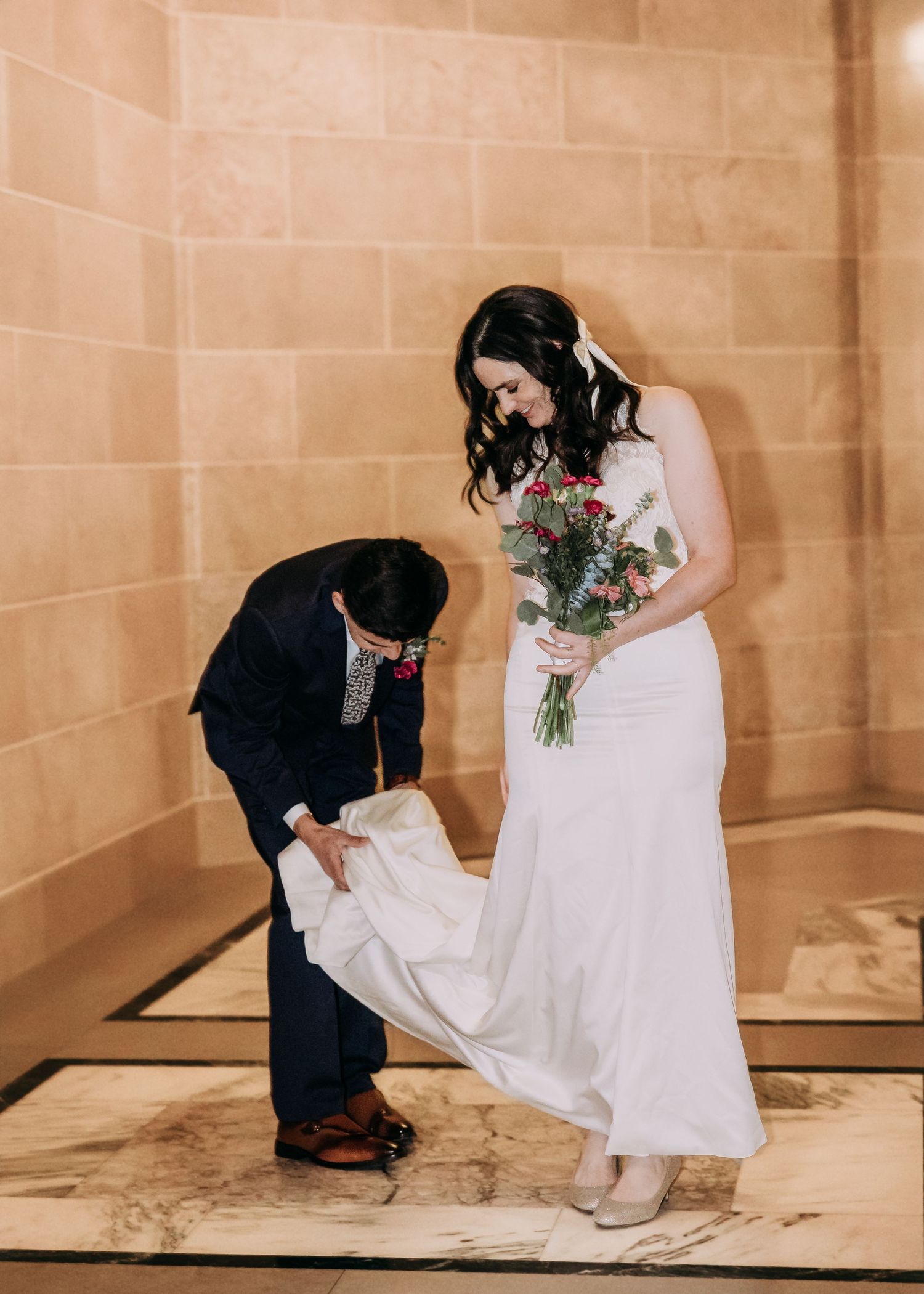 Groom carefully adjusts bride's white gown train while she smiles holding colorful bouquet.