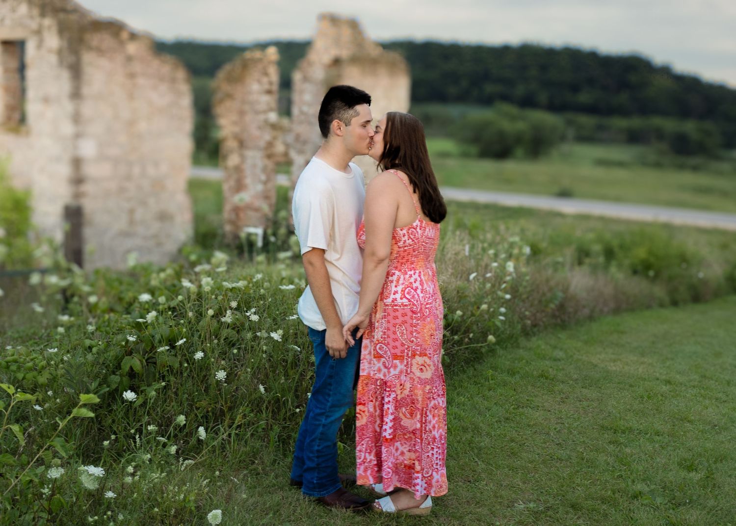 Couple sharing a kiss near rustic stone ruins surrounded by wildflowers and lush greenery.