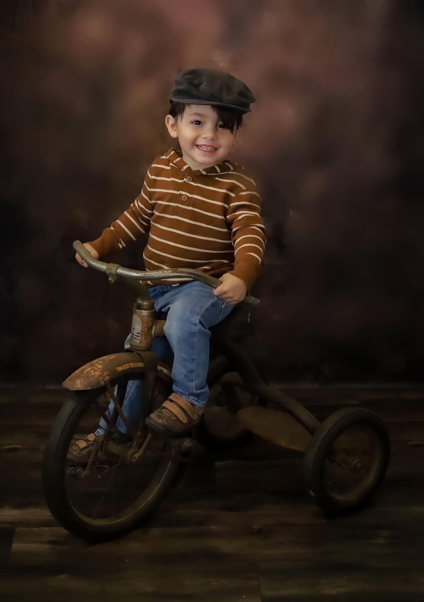 Smiling boy in striped brown sweater and newsboy cap rides vintage tricycle in studio.