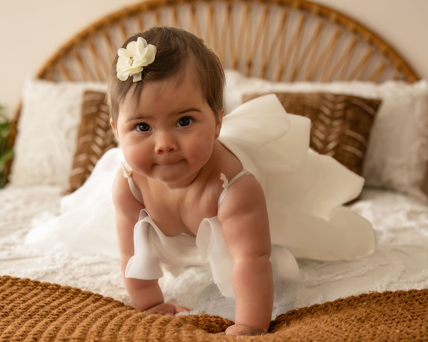 A cute baby girl in a white tulle dress and flower headband crawls on a textured golden blanket.