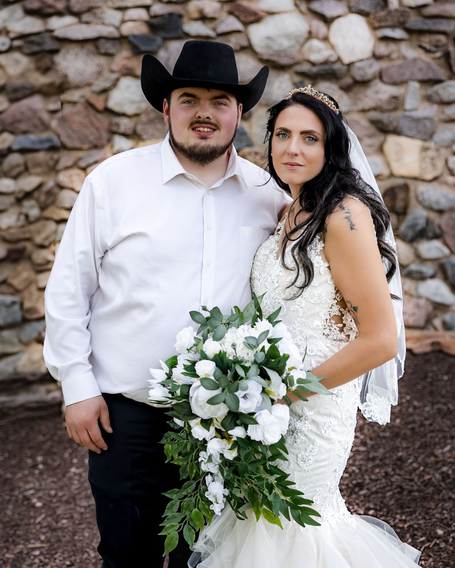Bride in lace mermaid gown holding white floral bouquet stands with groom in black cowboy hat and white shirt.
