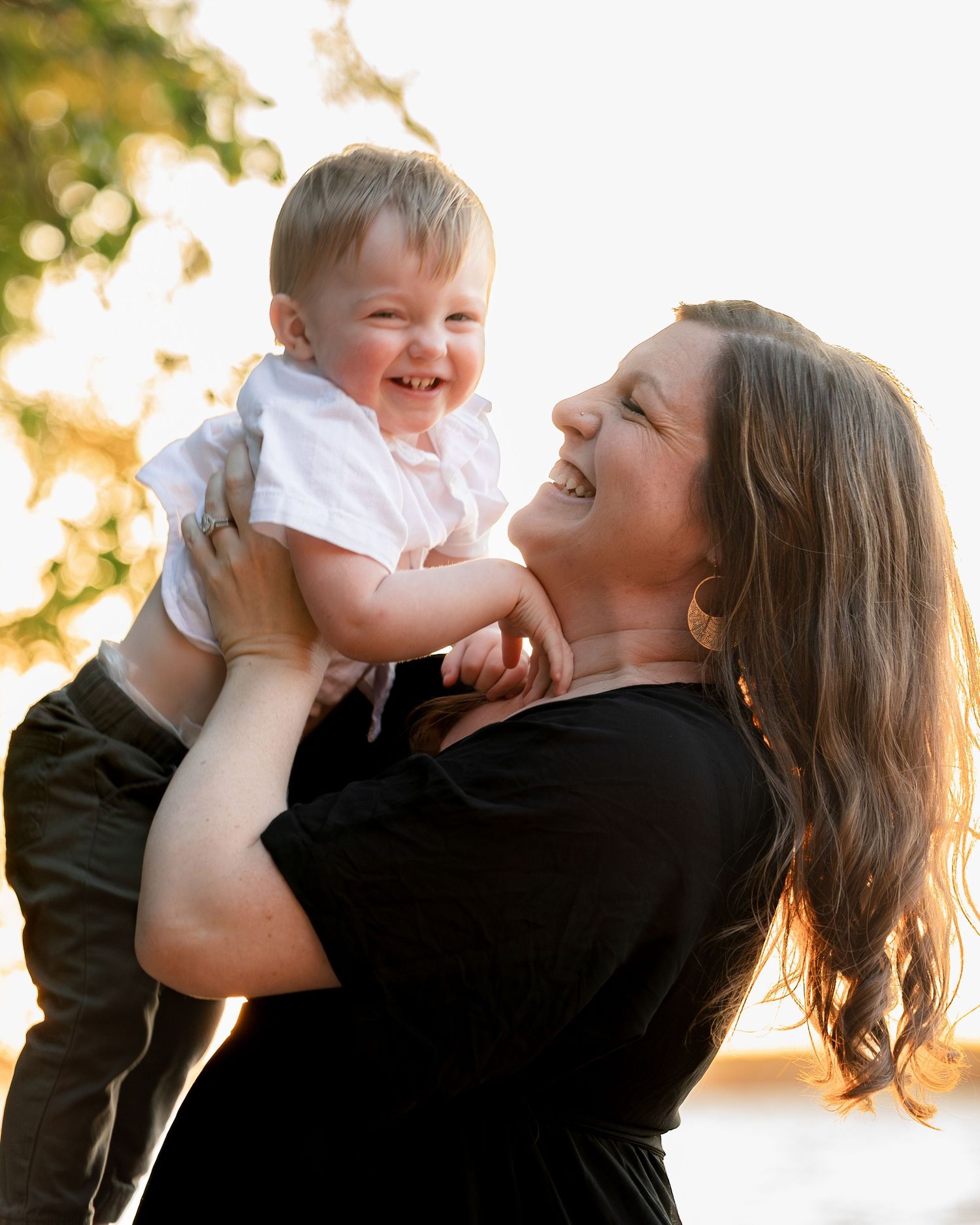 A joyful mother lifts her laughing baby boy outdoors in warm golden sunlight, both sharing a happy moment.