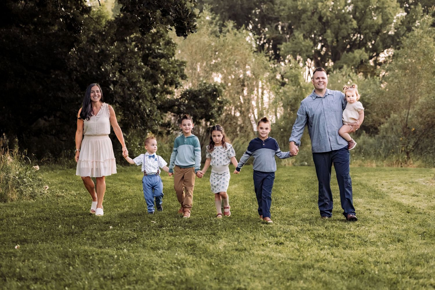 Family of seven walking hand-in-hand through a lush green park, parents with five young children in a candid outdoor portrait.