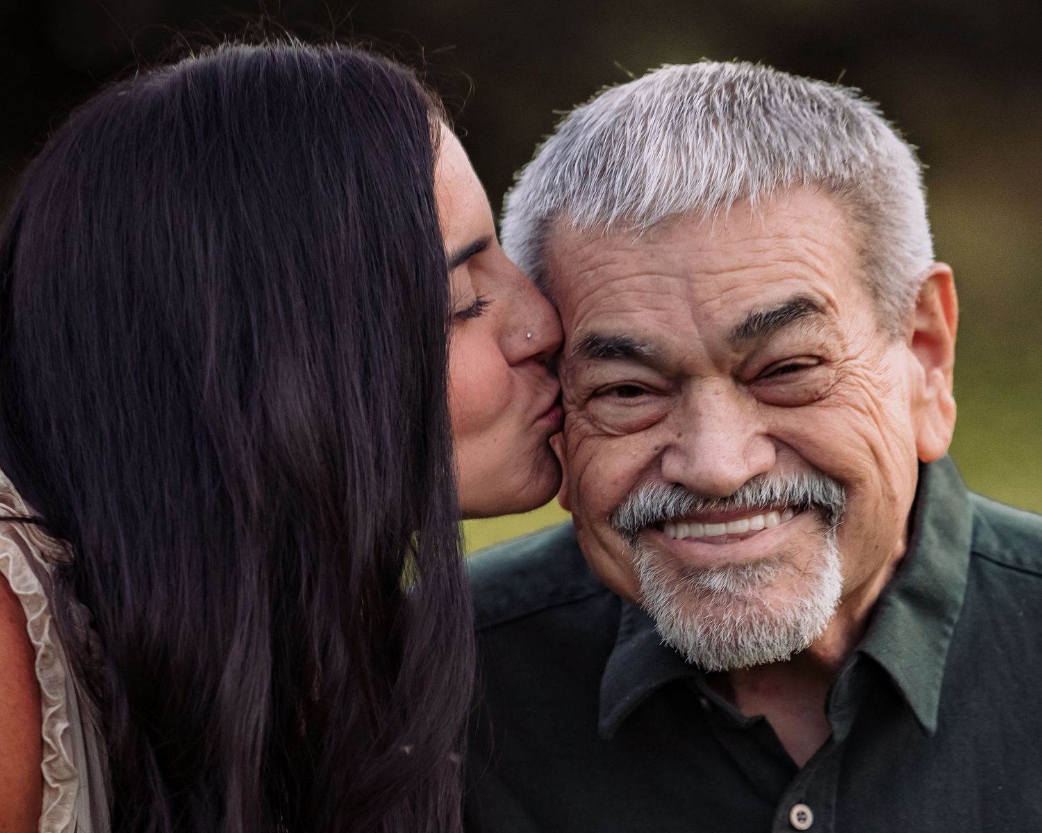 A young woman with dark hair kisses the cheek of a smiling older Hispanic man with gray hair and mustache.