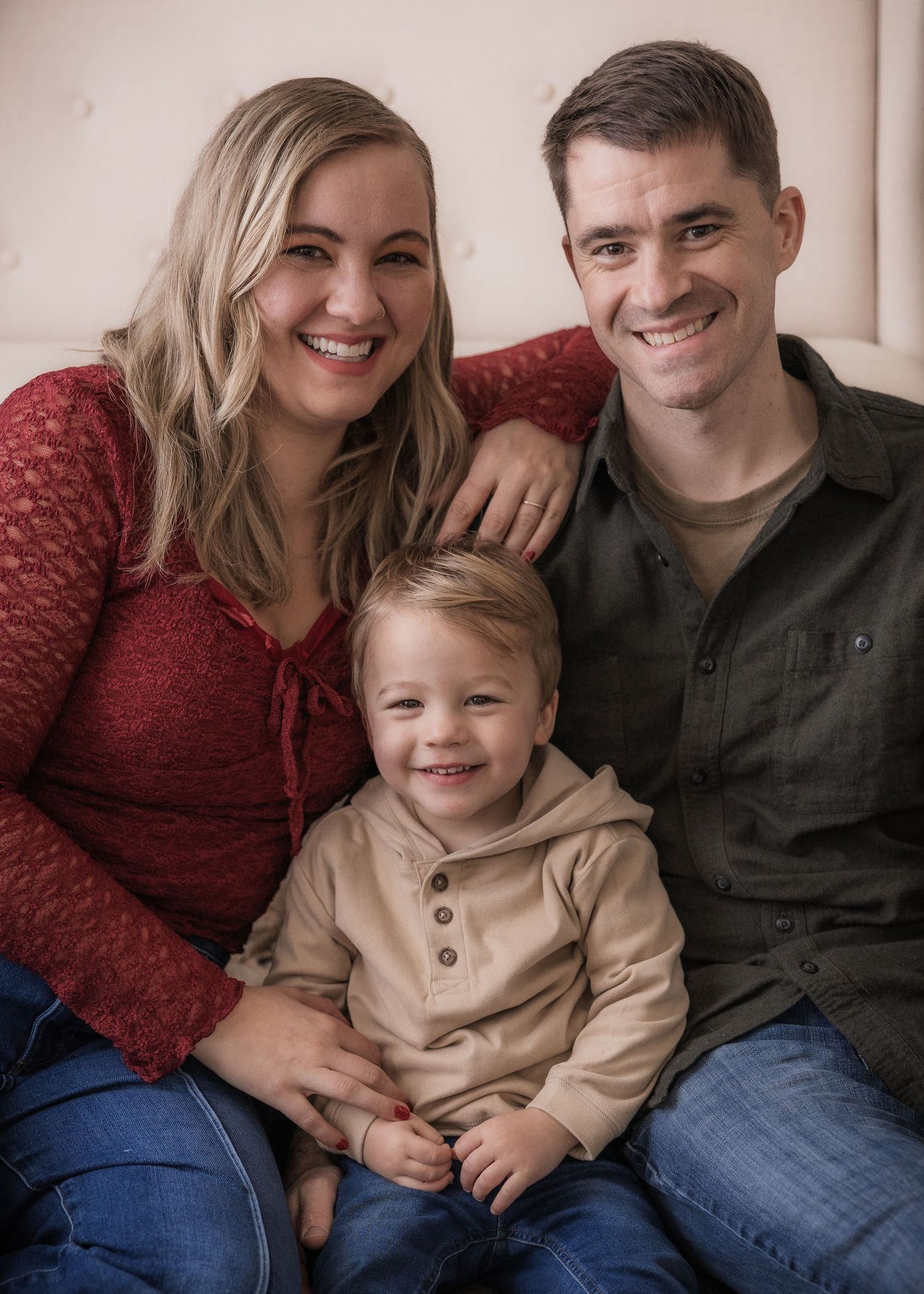 Happy family portrait of smiling parents with toddler boy sitting together on a couch in casual clothing.