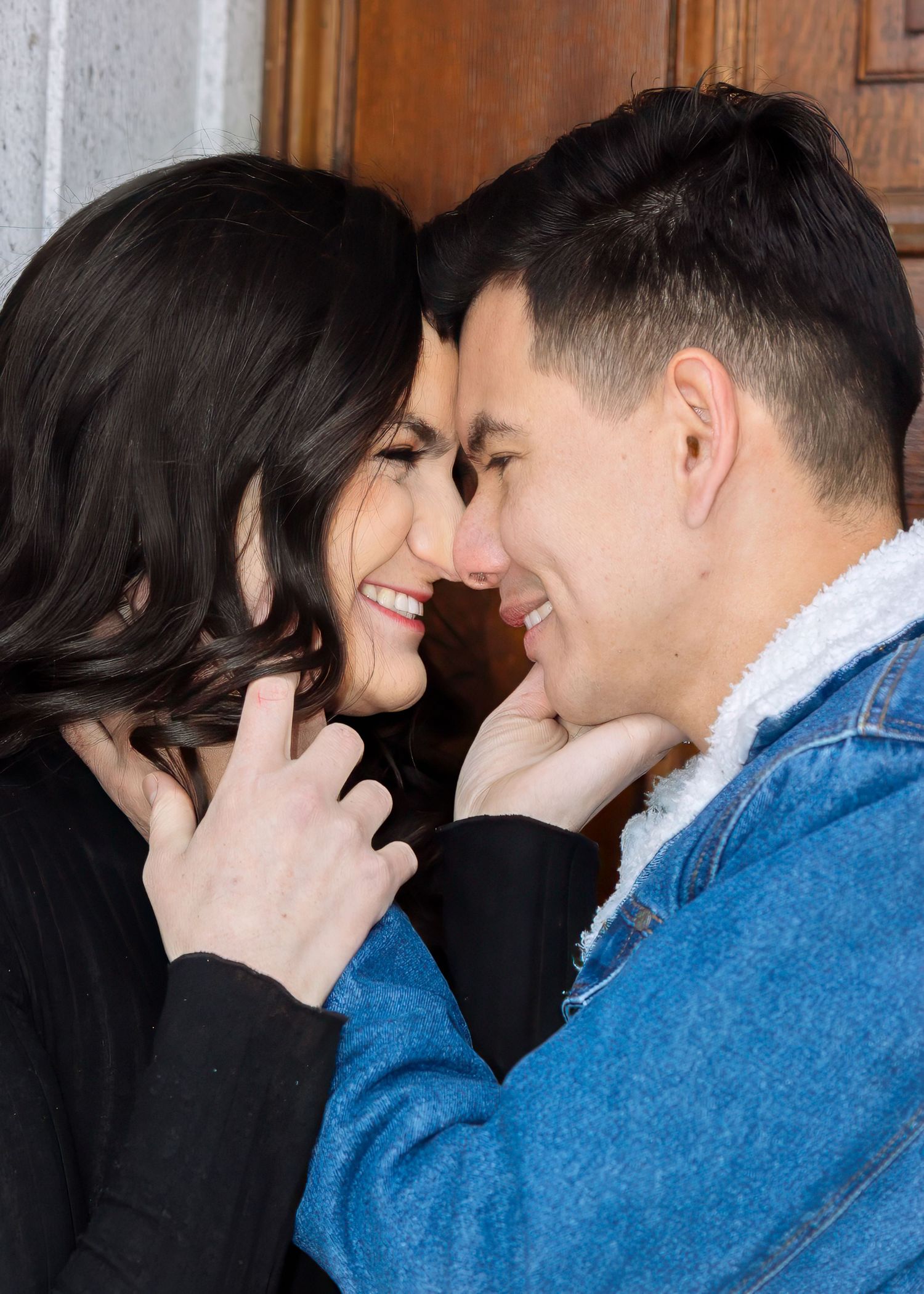 Happy couple shares an intimate moment near a wooden door, woman in black top touching mans chin tenderly.