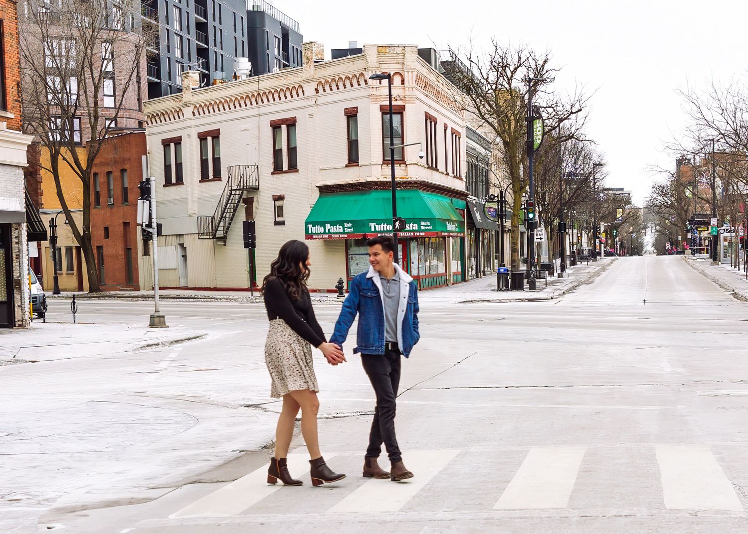 Couple holding hands crossing a city street intersection with a green awning storefront behind them.