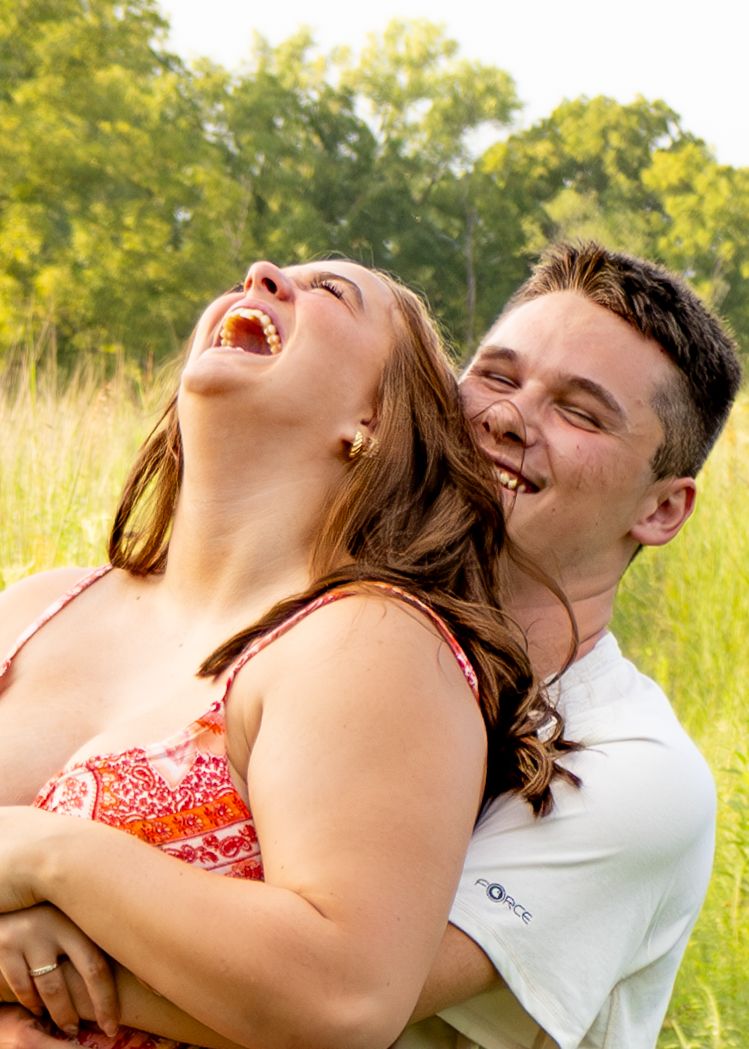 Joyful couple laughing together in a grassy field, woman in red floral dress throwing head back.