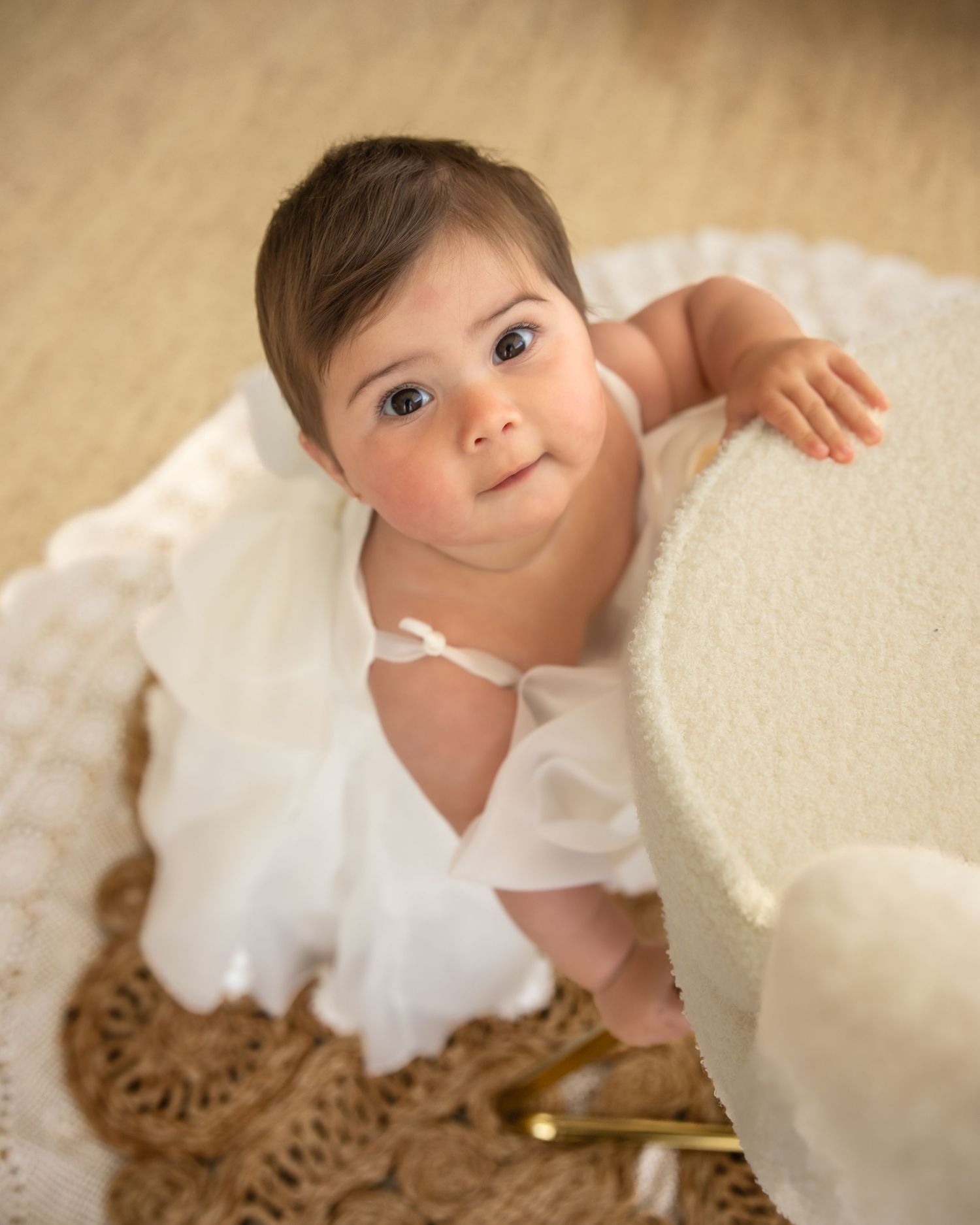 Adorable baby in white dress looks up curiously while leaning against a white chair.