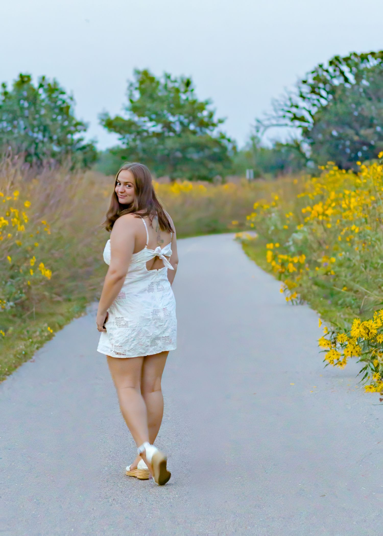 A young woman in a white mini dress walks along a path lined with yellow wildflowers.