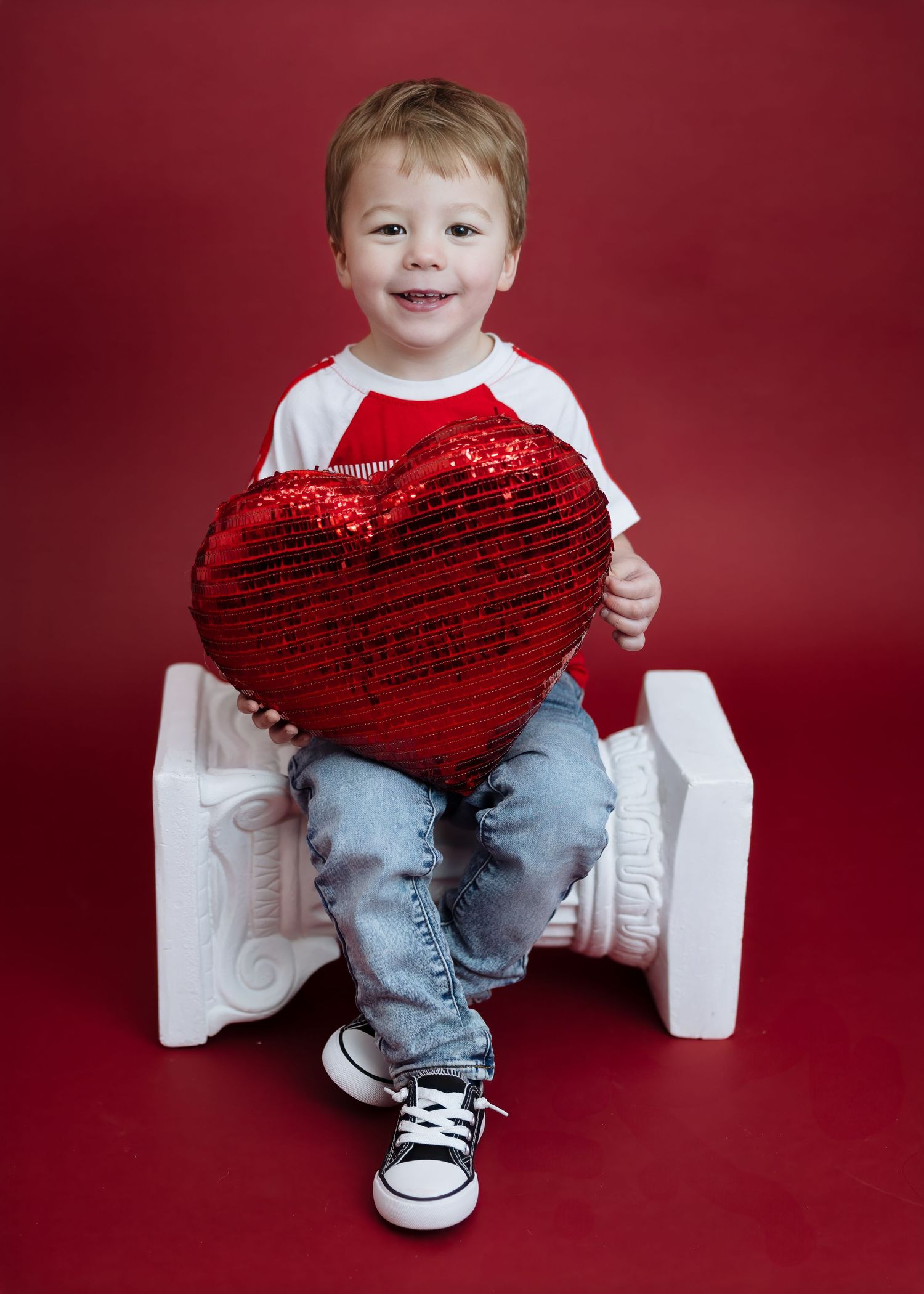 Smiling toddler holds red sequin heart pillow seated on white pedestal.