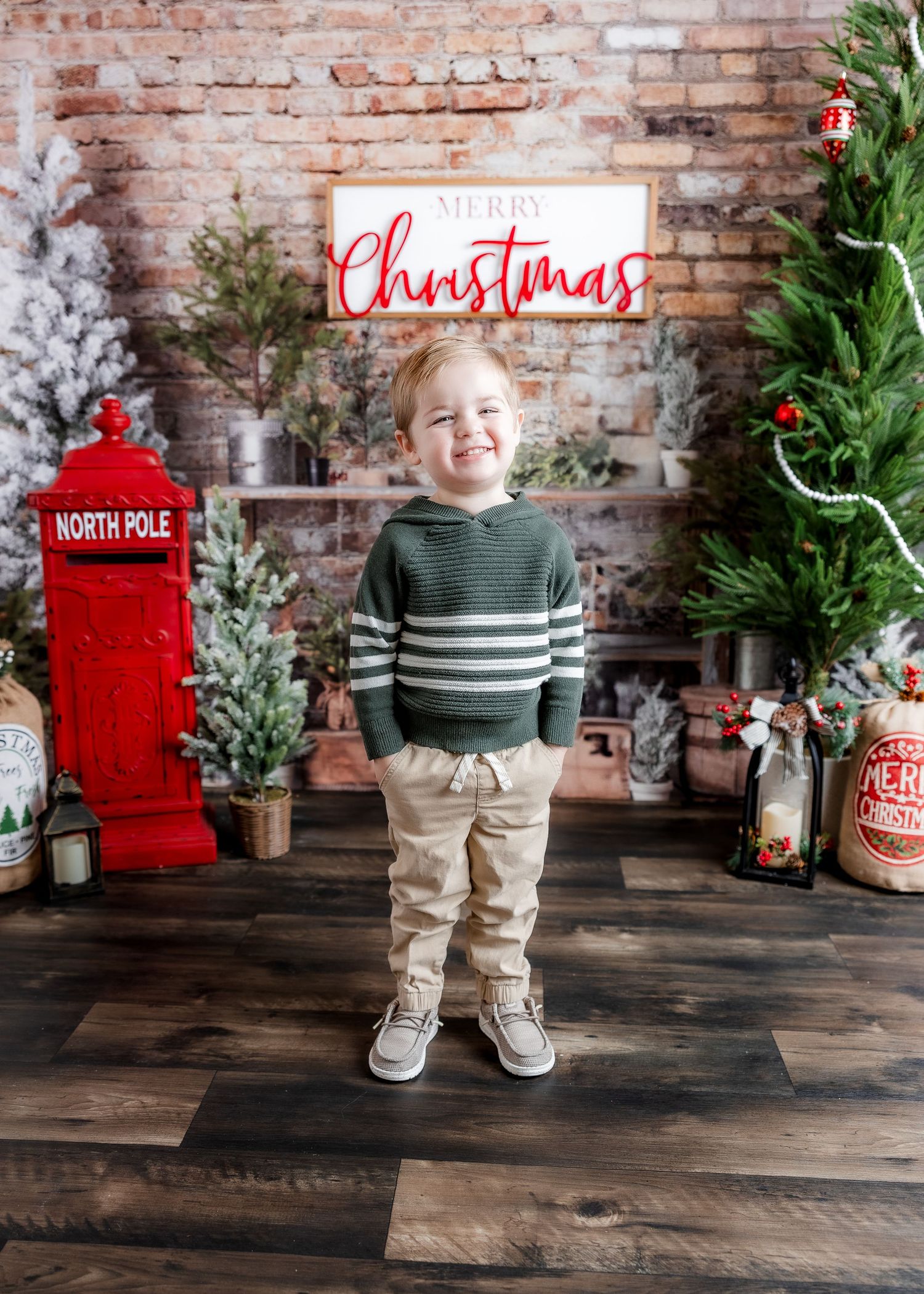 A cheerful boy in a green striped sweater stands smiling in a festive Christmas photo studio with holiday decorations.